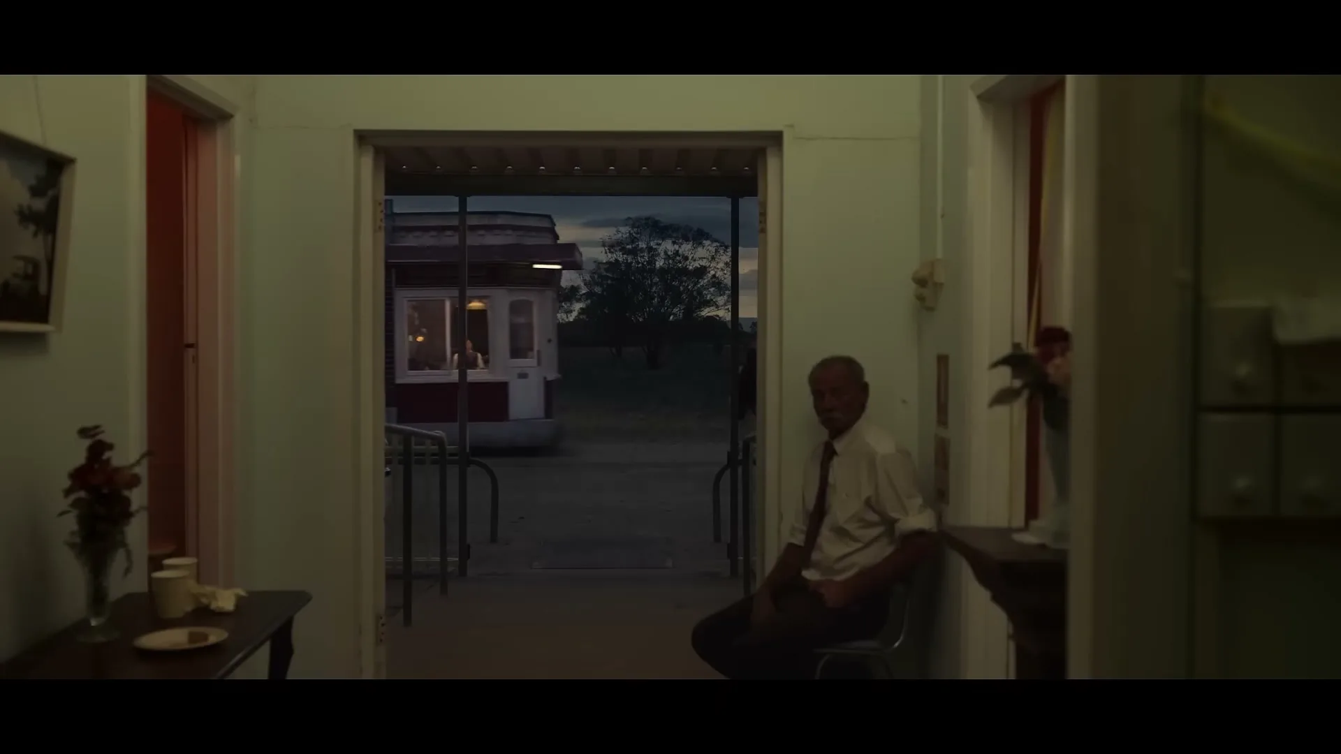 An older man sits in a dimly lit interior, looking at the camera, while a ticket booth is visible through a doorway in the distance at dusk.