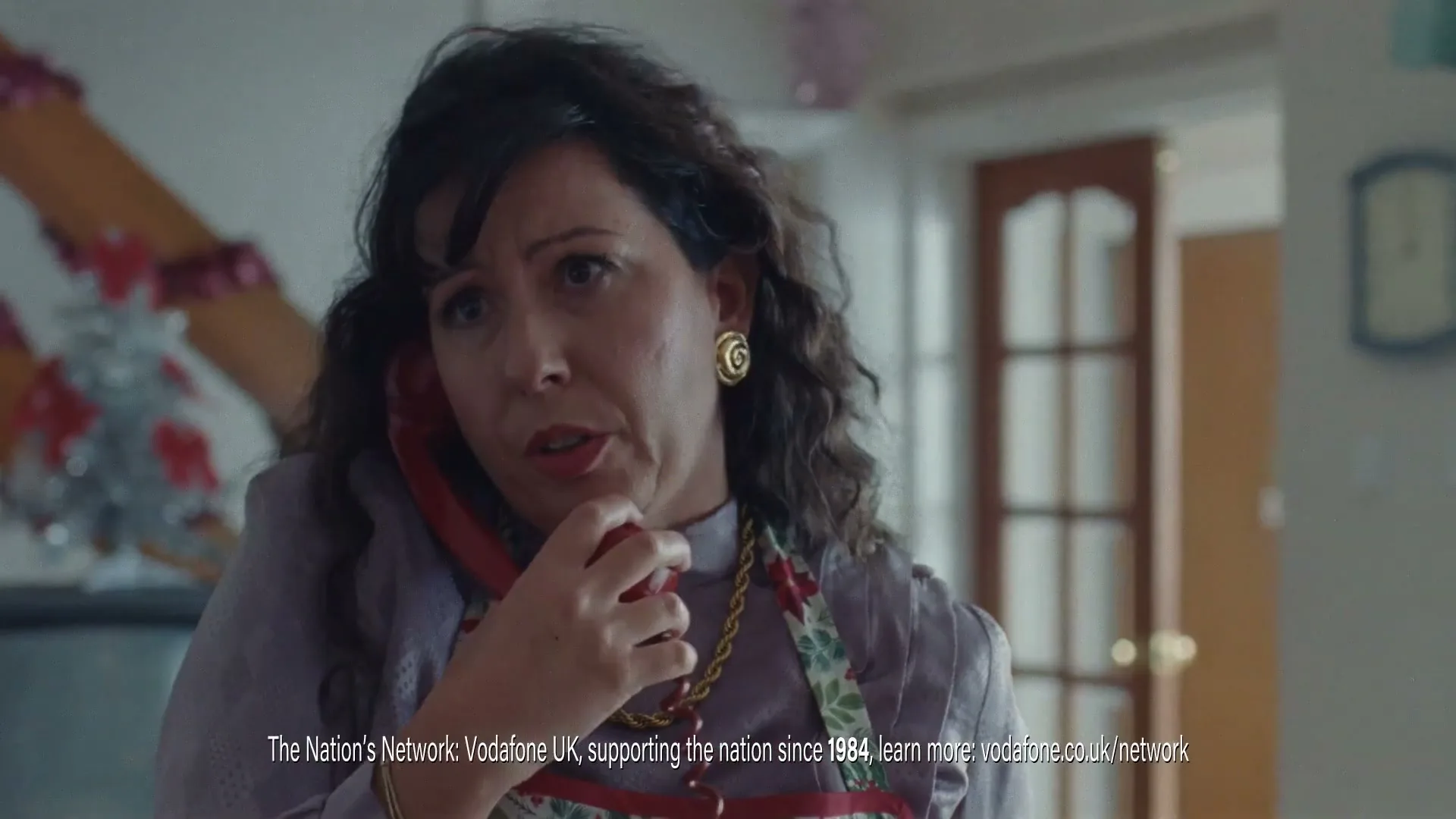 A woman in her 40s, wearing a purple blouse and a floral apron, talks on a red corded phone with a concerned expression in a decorated living room.