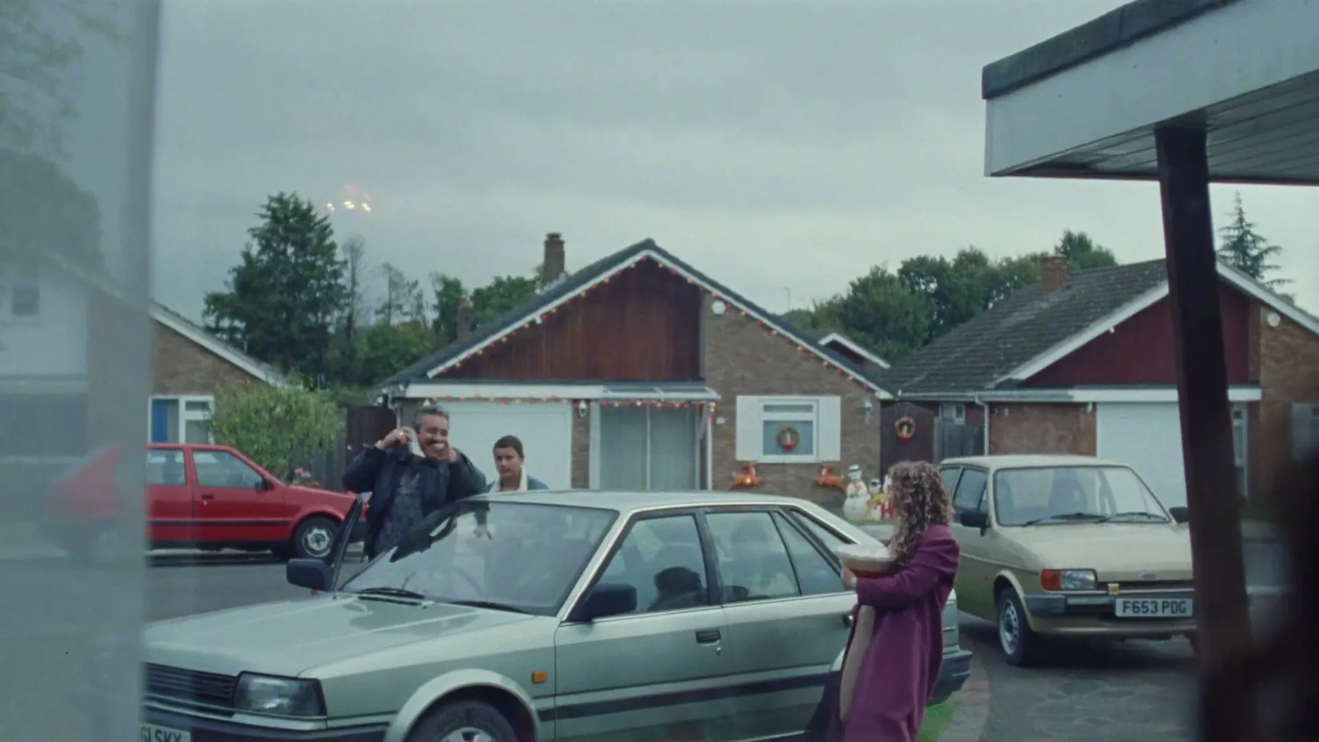 A family arrives at a suburban house decorated for Christmas, with a man talking on a phone and a woman carrying a bowl.
