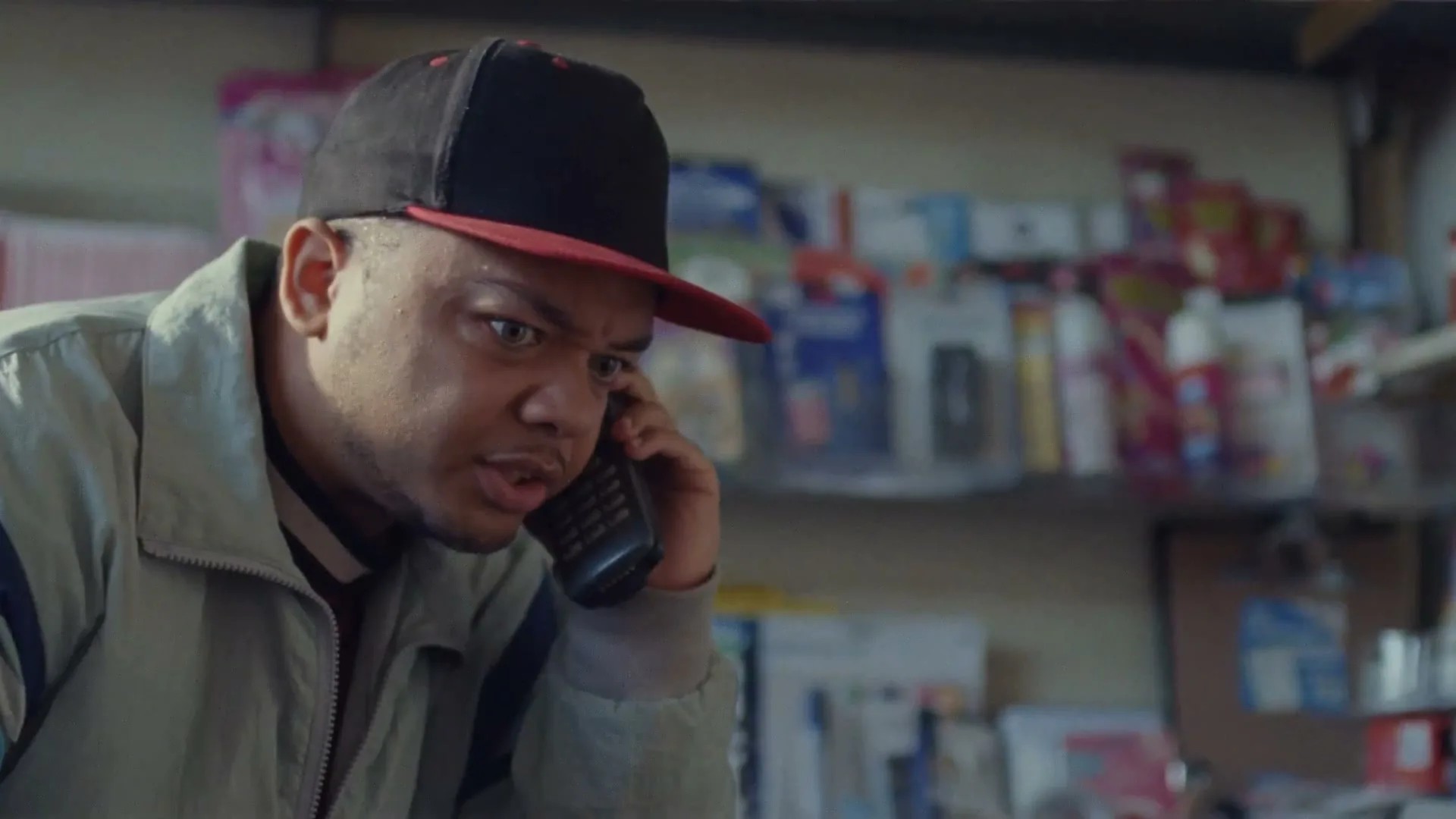 A young man in a cap and jacket talks on an old-fashioned mobile phone, looking confused in a cluttered shop.