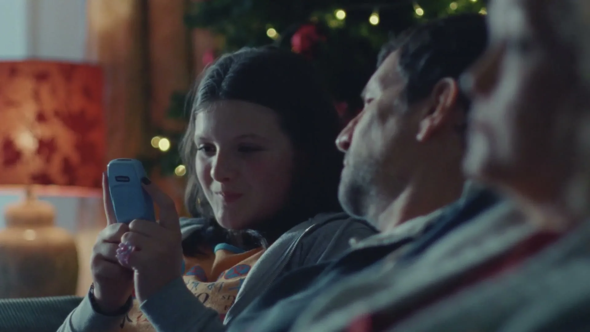 A young girl with dark hair sits on a couch, holding a light blue flip phone, looking at an older man next to her, with a Christmas tree in the background.