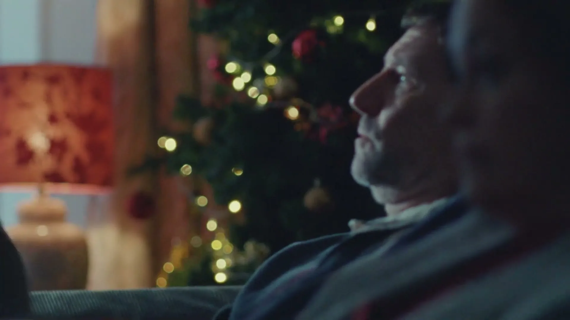 A young woman in a t-shirt and jacket looks at a man with a beard, who is looking up, as they sit on a couch in a living room with a Christmas tree.