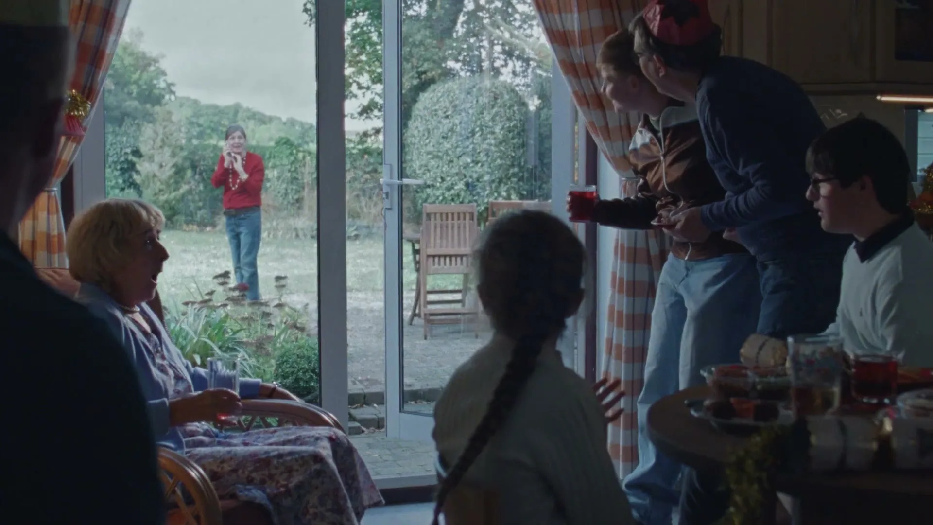A family gathers inside a home for Christmas, while a young boy stands outside in the garden, looking in through a glass door.