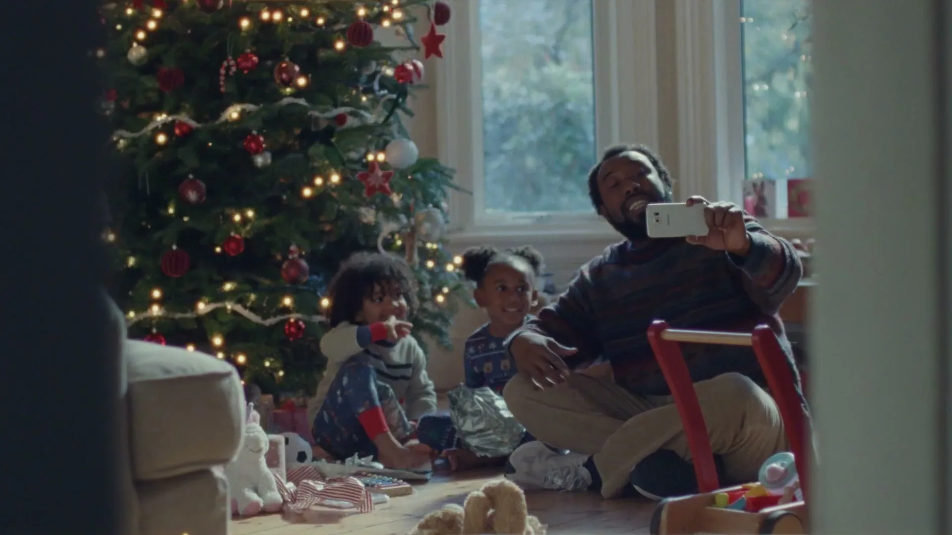 A father sits on the floor with his two young children, taking a selfie in front of a decorated Christmas tree in a cozy living room.