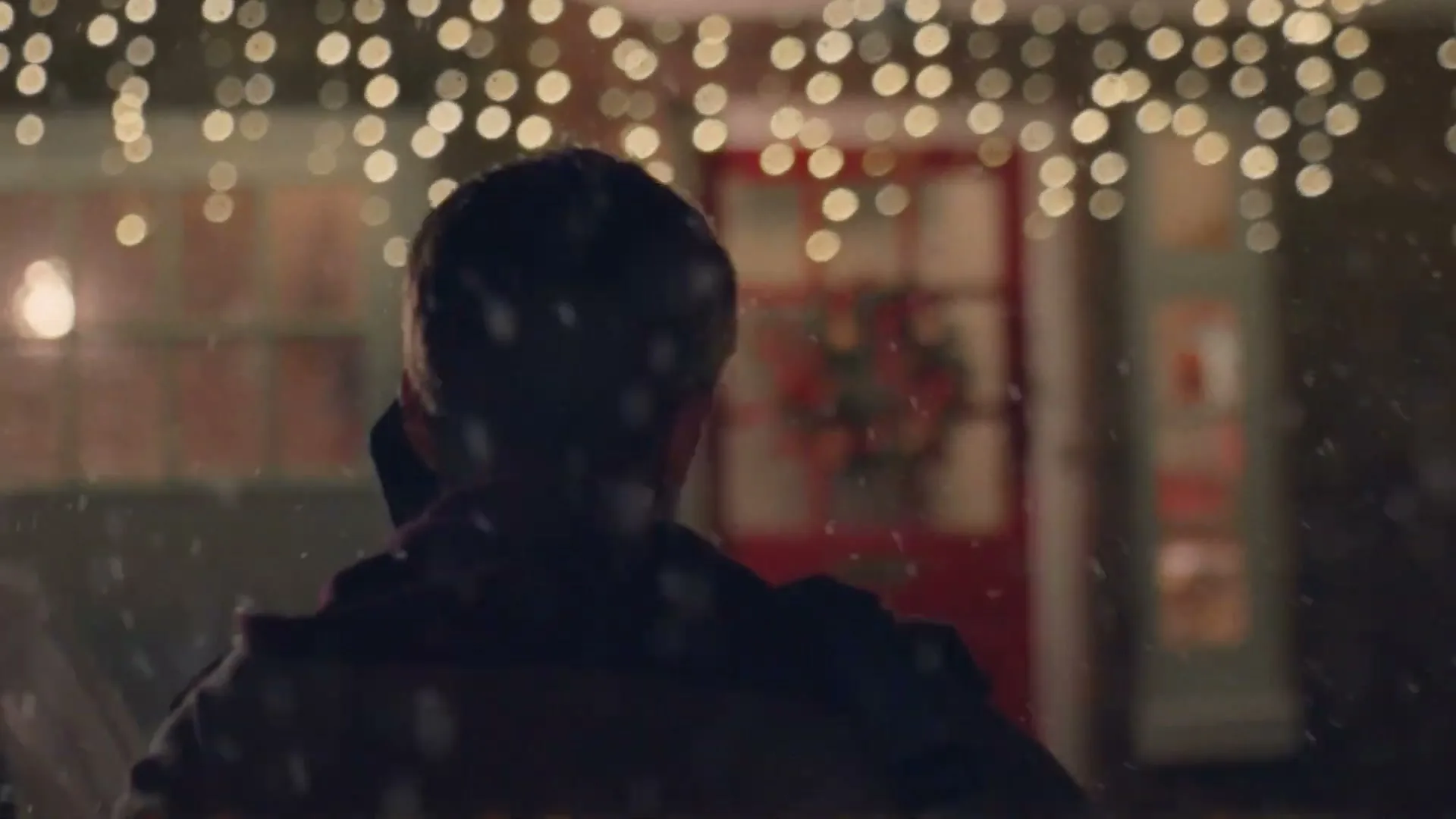 A man stands with his back to the camera, looking at a house decorated with Christmas lights and a wreath, as snow falls around him at night.