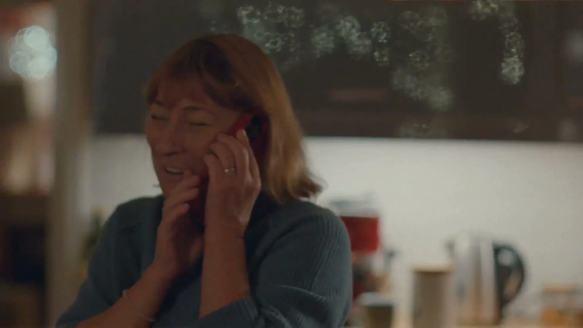 A middle-aged woman with shoulder-length brown hair, wearing a blue sweater, smiles while holding a red smartphone to her ear in a cozy kitchen.