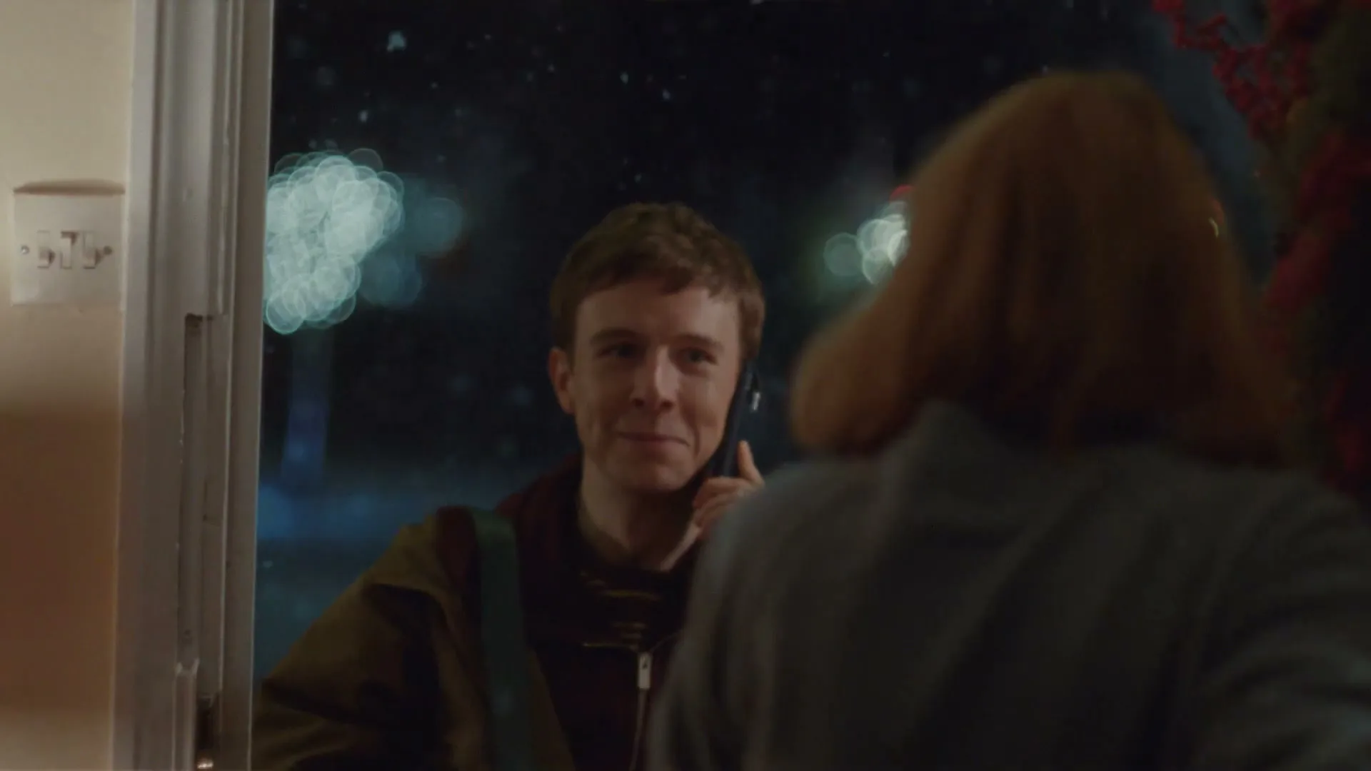 A young man with a phone to his ear smiles at a woman with red hair, who is seen from behind, standing in a doorway at night.