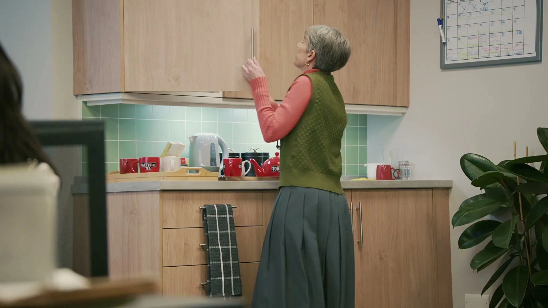 A senior woman with short grey hair, wearing a green vest and pink long-sleeve shirt, opens a kitchen cabinet in an office kitchen.