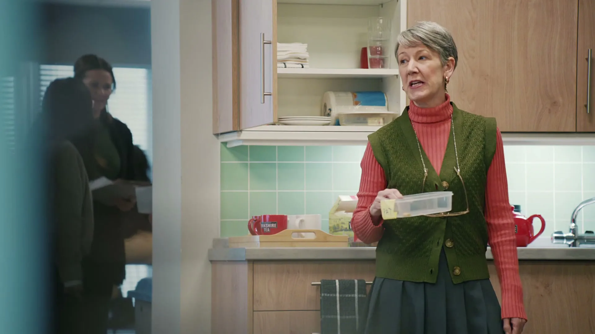 A grey-haired woman in a green vest and pink turtleneck holds a clear container and looks at the camera in an office kitchen.