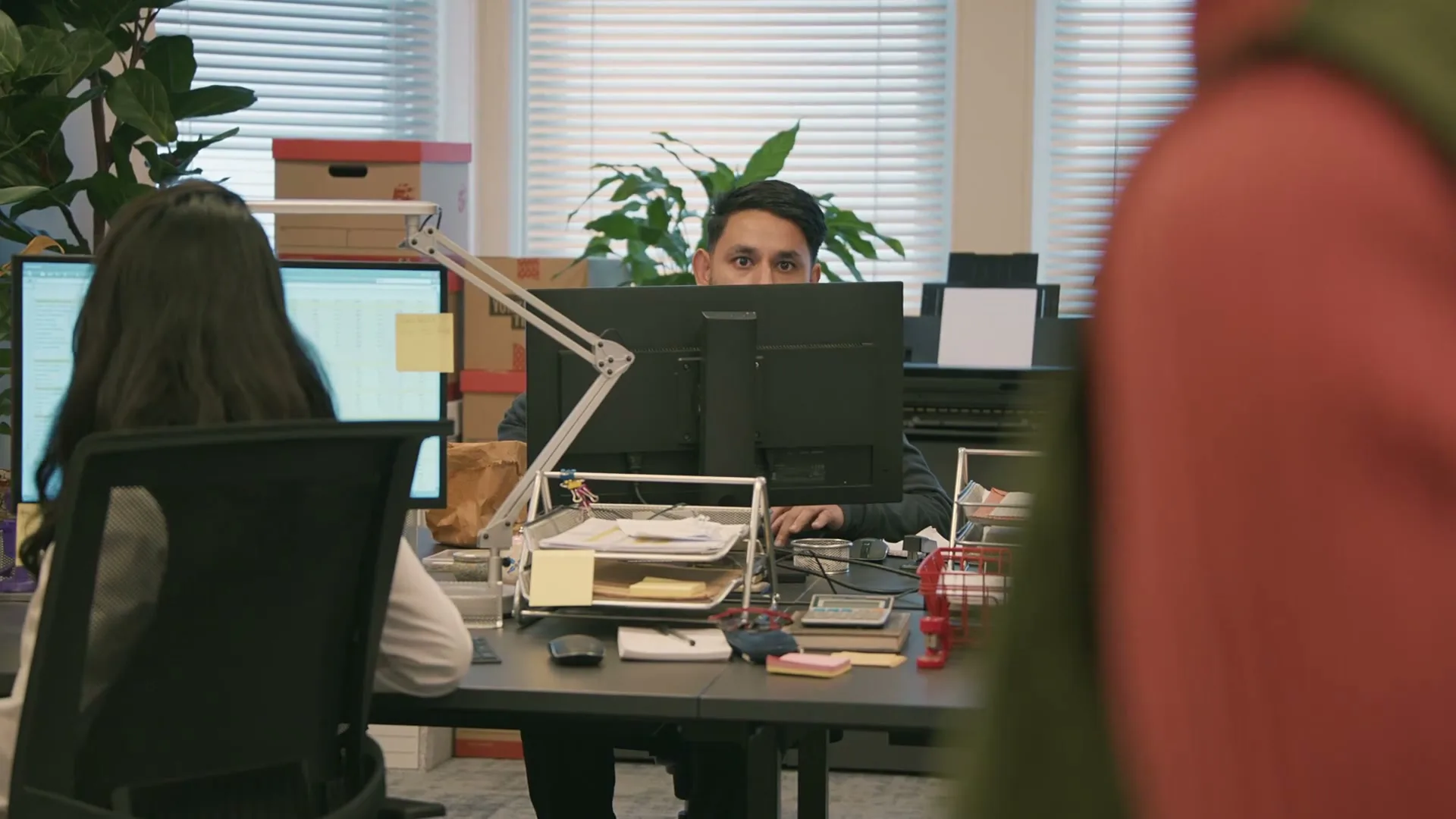 A man with dark hair peeks over his computer monitor, looking intently at something off-screen in a modern office setting.