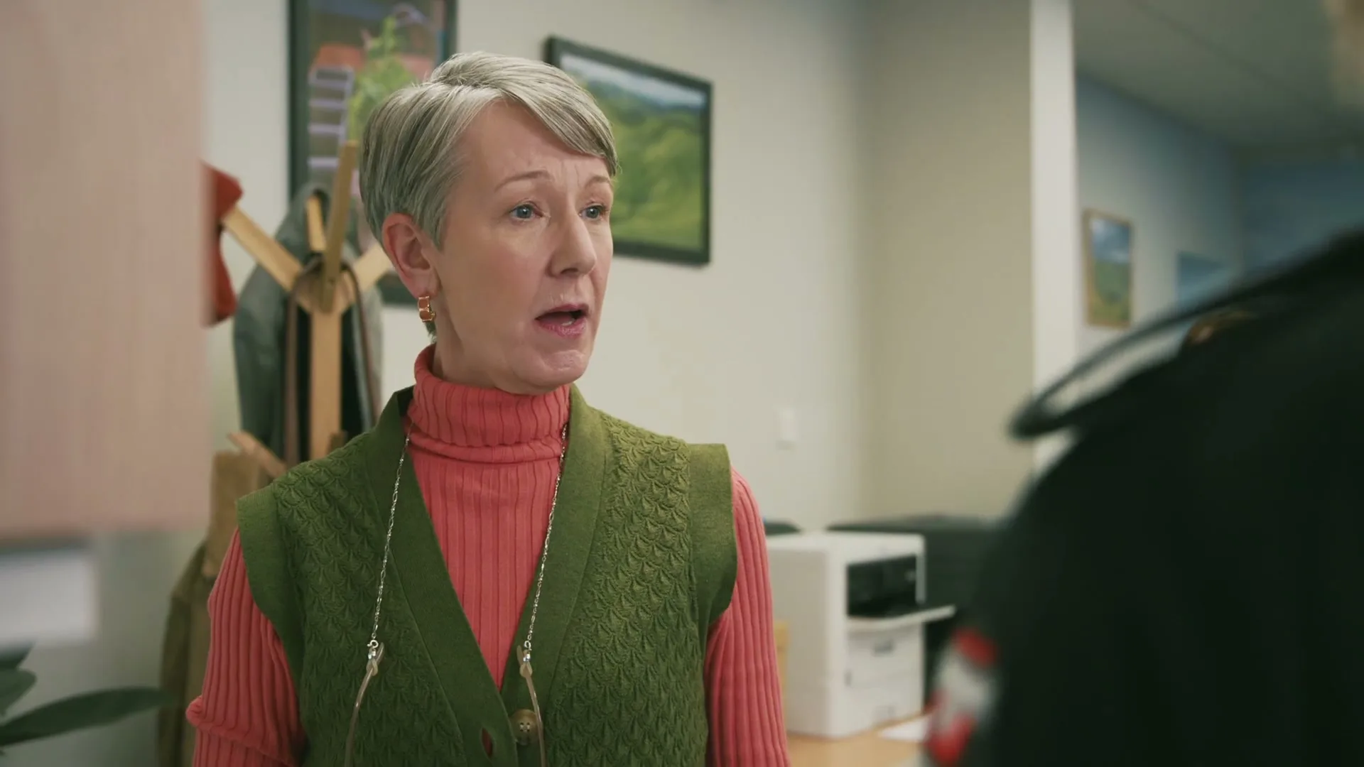 A middle-aged woman with short grey hair and a green vest over a pink turtleneck talks to someone off-screen in an office.