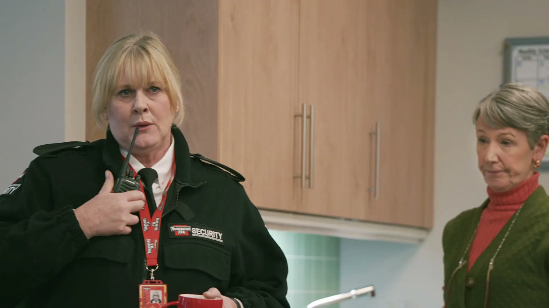 A female security guard in a black uniform holds a red mug and a walkie-talkie, speaking into it while another woman stands beside her in an office kitchen.