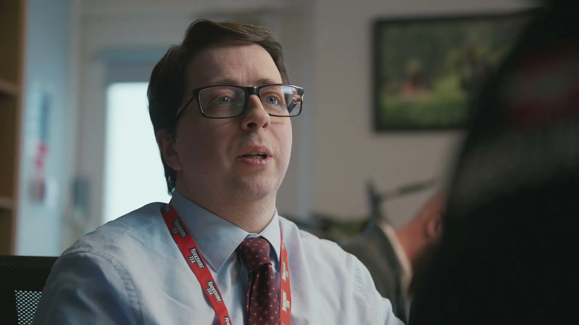 A man in his 30s, wearing glasses and a shirt with a tie, speaks to someone off-camera in an office setting.