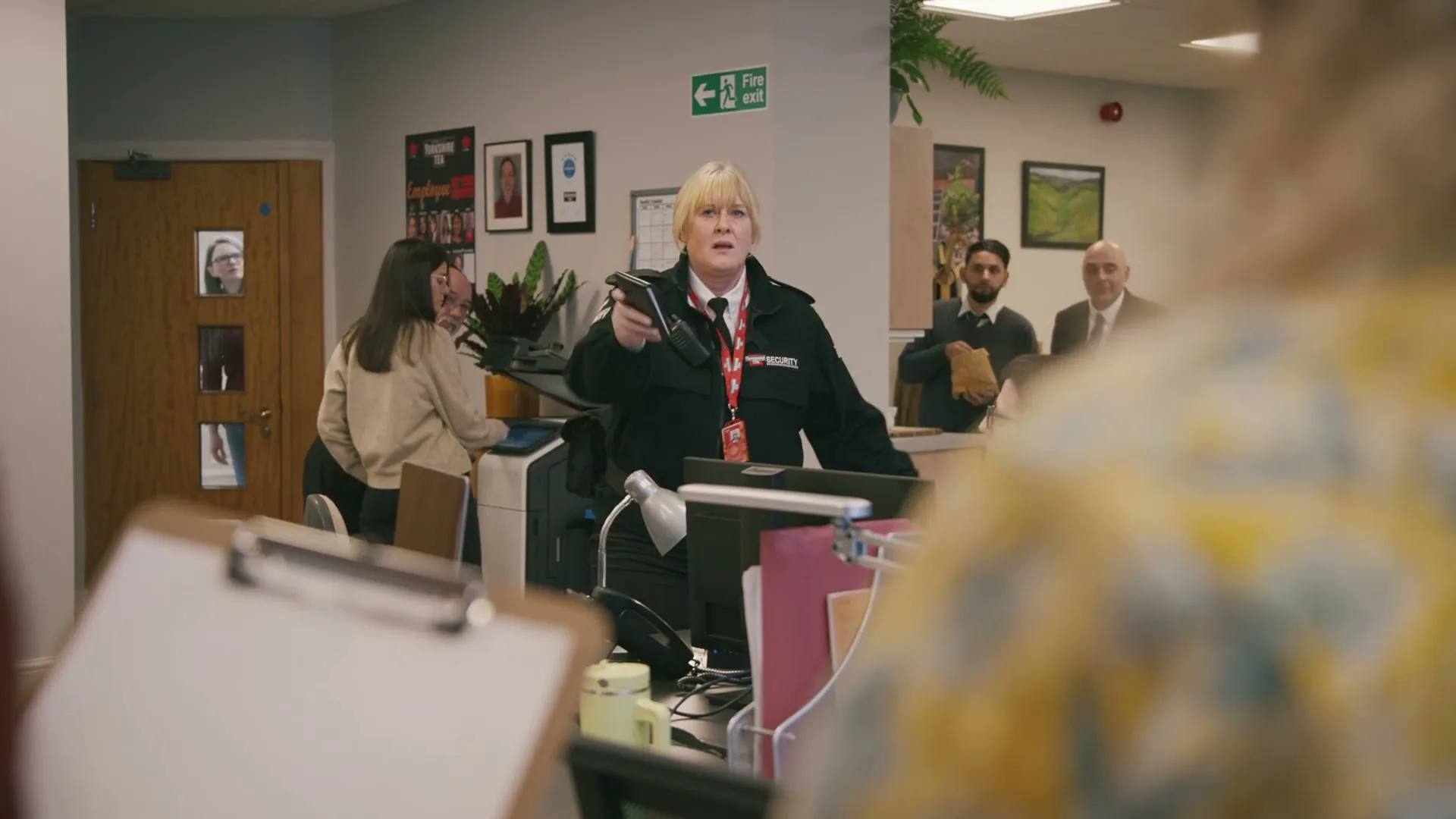 A female security guard in a black uniform points a walkie-talkie at someone off-screen in a busy office, while other employees look on.