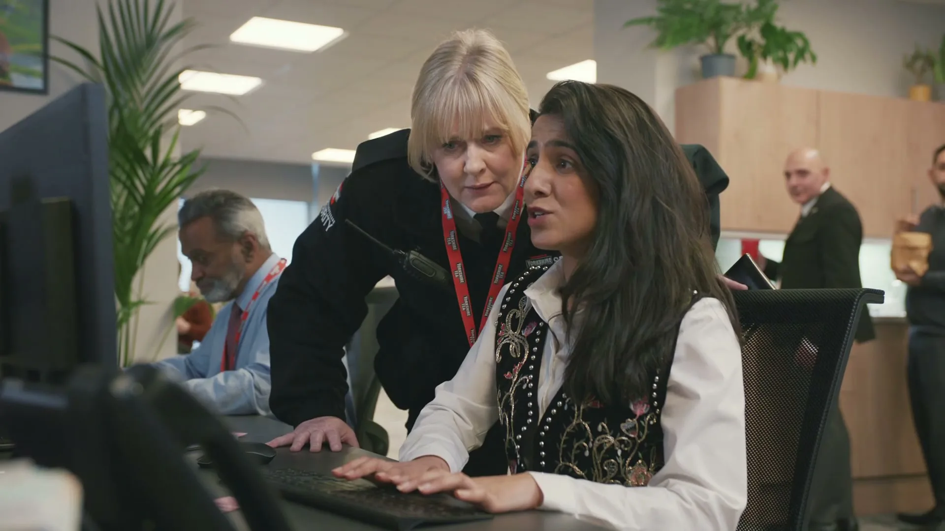 A female police officer leans over a young woman working at a computer in a busy office, looking at her screen with a concerned expression.