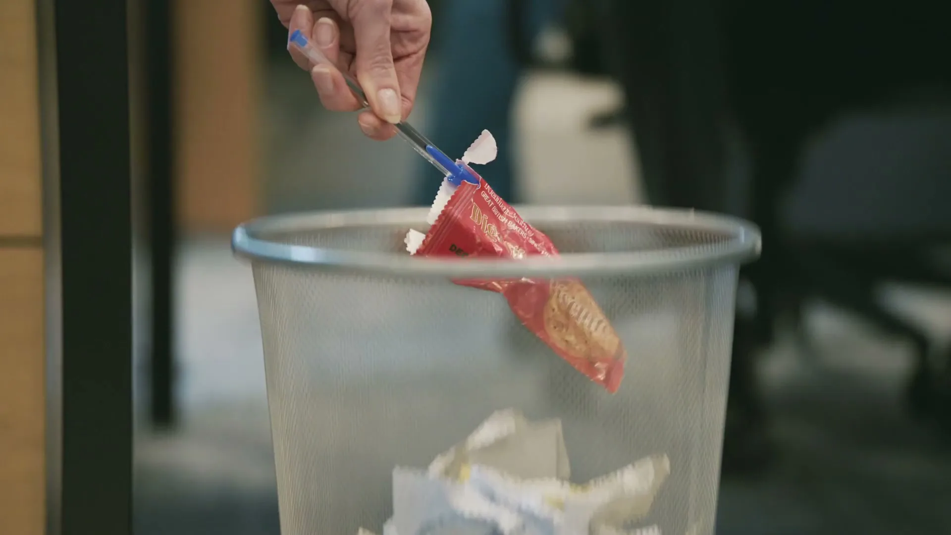 A person's hand uses a pen to retrieve a red wrapper from a metal mesh trash can in an office setting.