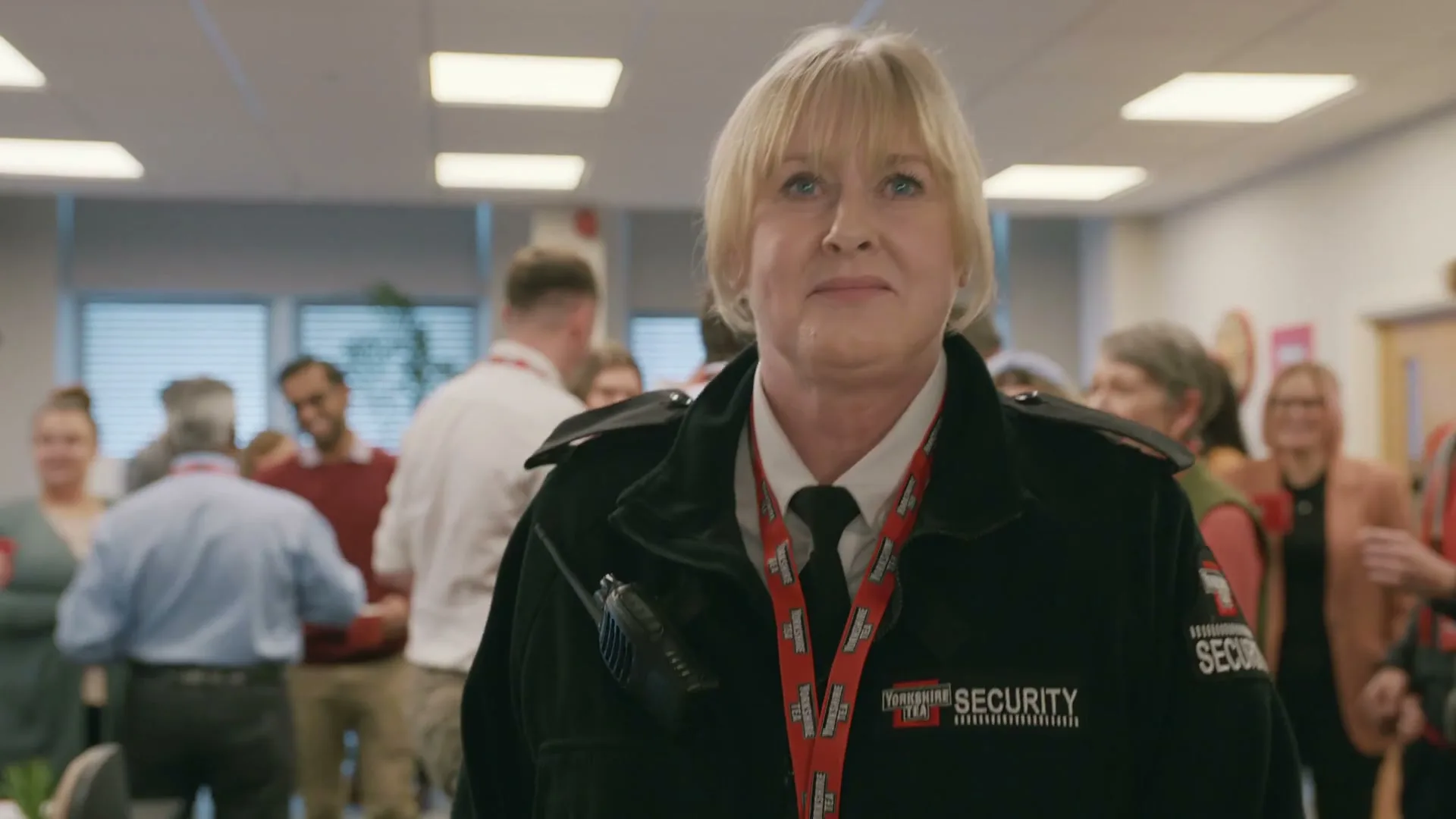 A female security guard with blonde hair walks through a crowded office, looking determined as people clap and cheer around her.