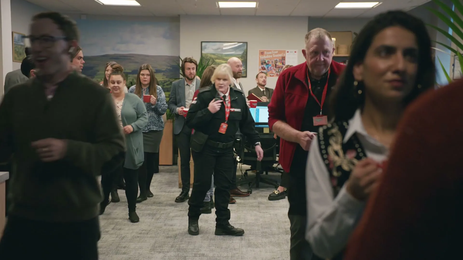 A female security guard in a black uniform walks through a crowded office, speaking into a walkie-talkie, as people hold red mugs and watch her.