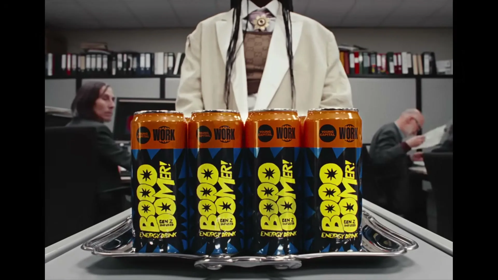A person in a white suit holds a tray of "Boomer!" energy drinks in an office setting, with other office workers in the background.