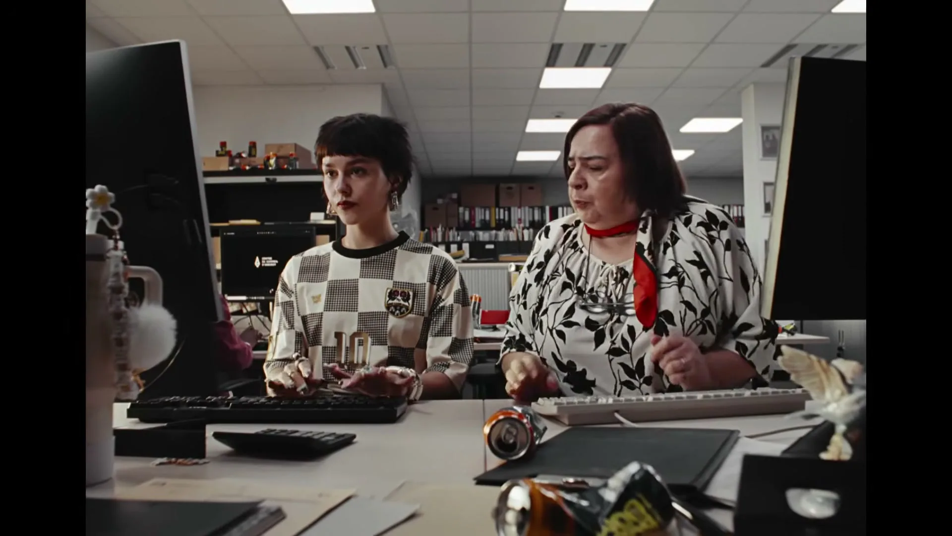 Two women sit at desks in an office, typing on computers; one sips from a can while the other focuses intently on her screen.
