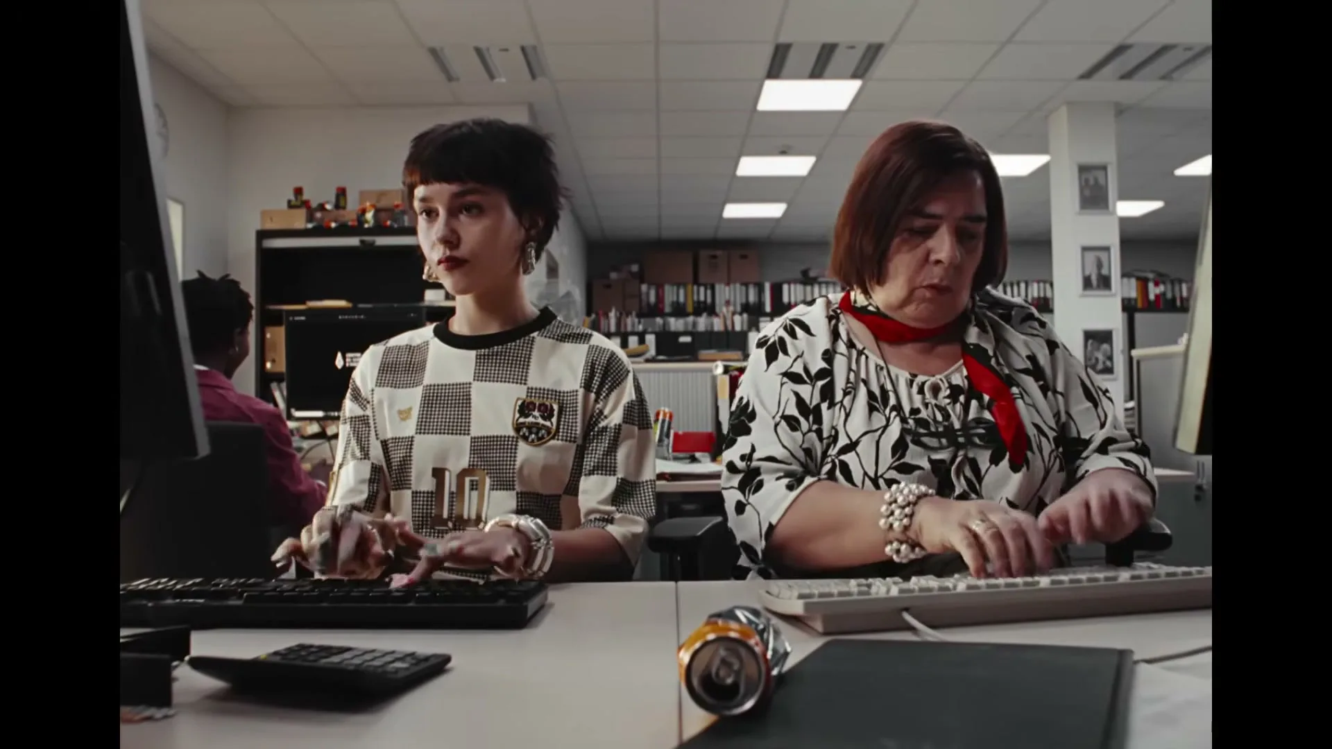 Two women, one young and one older, sit side-by-side at desks in an office, typing on keyboards.