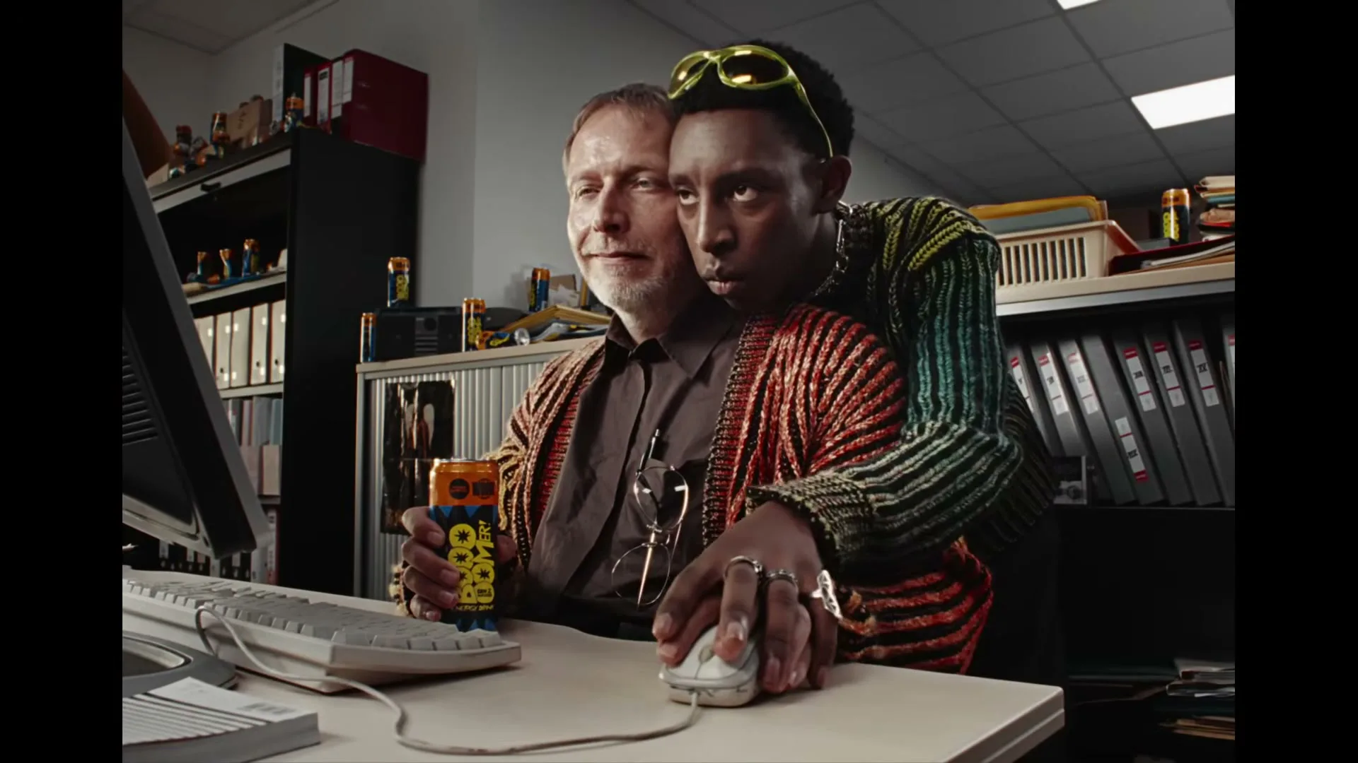 A young man with sunglasses and a colorful cardigan embraces an older man from behind, both looking at a computer screen in an office.