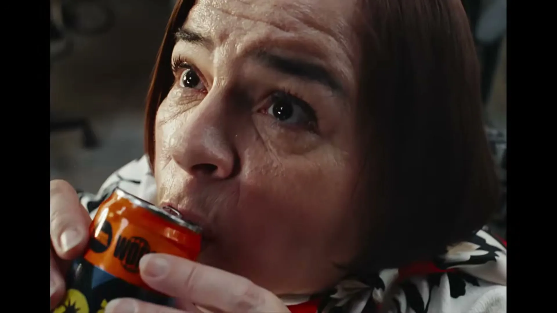 A woman with short brown hair and a patterned shirt drinks from an orange soda can, looking up with wide eyes in an interior setting.
