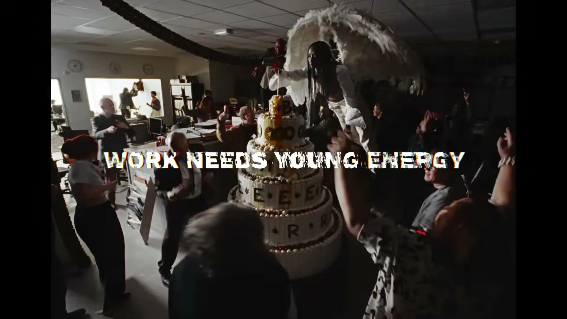 A large office party with many people dancing around a giant cake, while a person with angel wings stands atop the cake, and text overlays the scene.