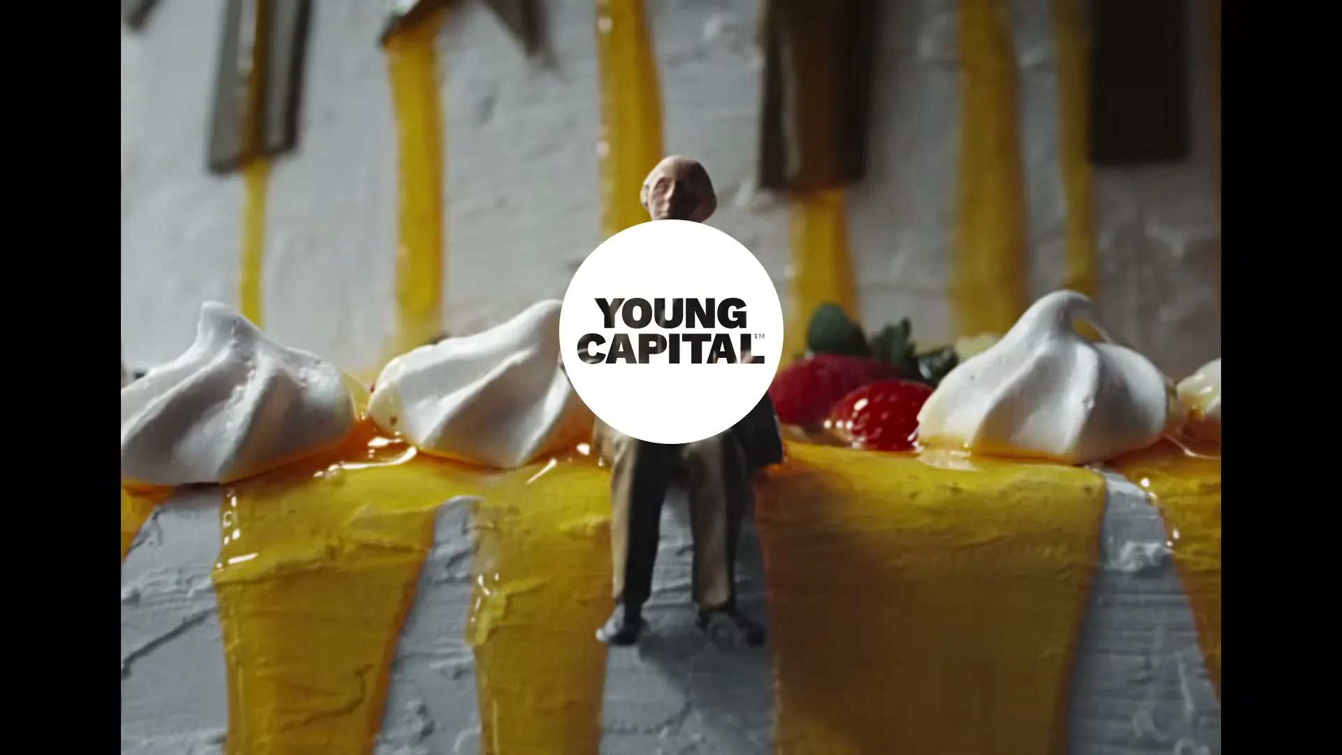 A miniature male figure stands on a cake covered in white frosting, yellow glaze, meringue, and strawberries, with the Young Capital logo superimposed.