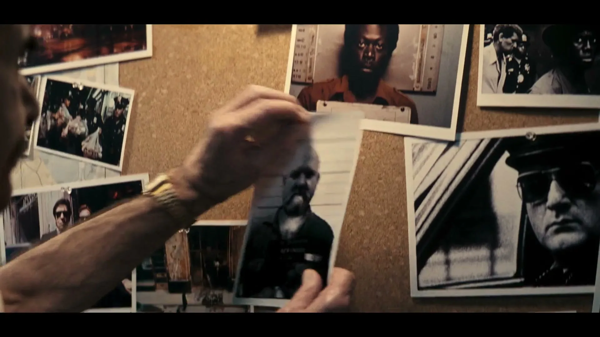 A man's arm with a gold watch pins a mugshot of a bald, bearded man onto a cork board covered with other crime scene photos and documents.