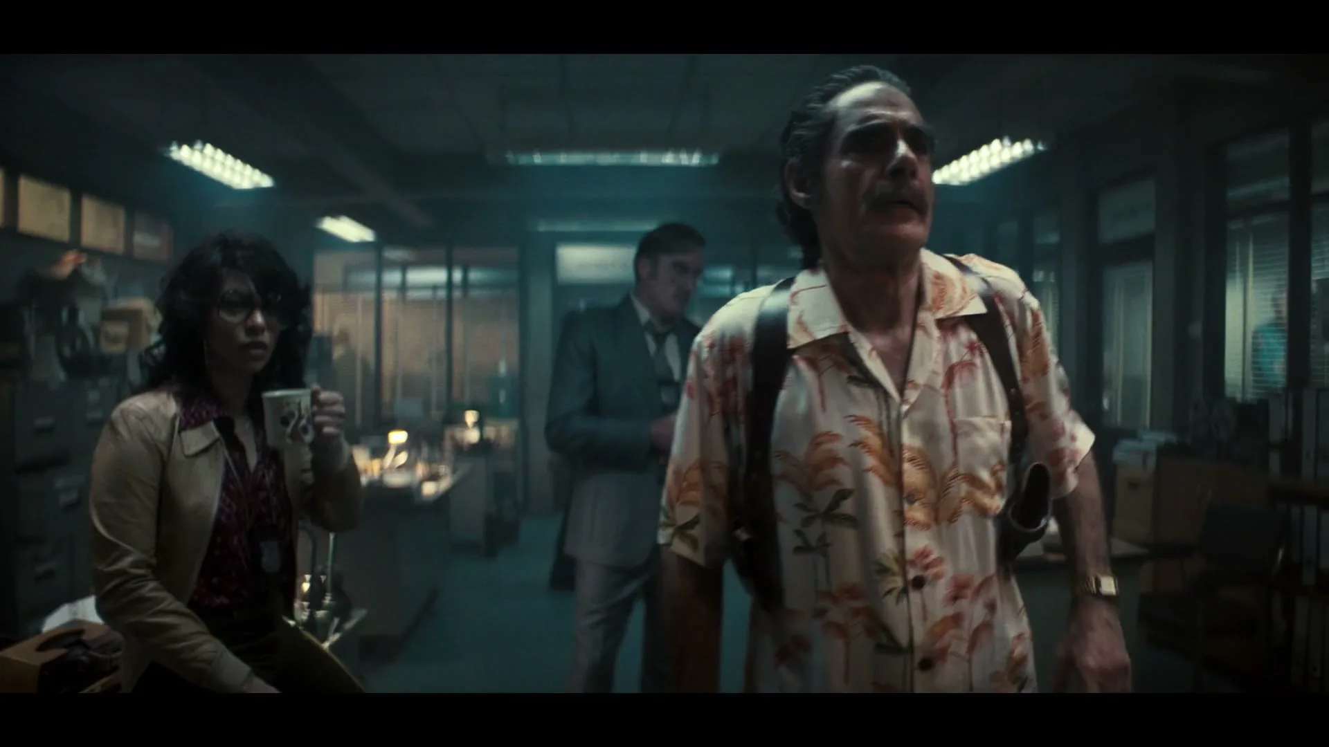 A male detective in a Hawaiian shirt and a female detective with a coffee mug look around a dimly lit, messy office with other people in the background.