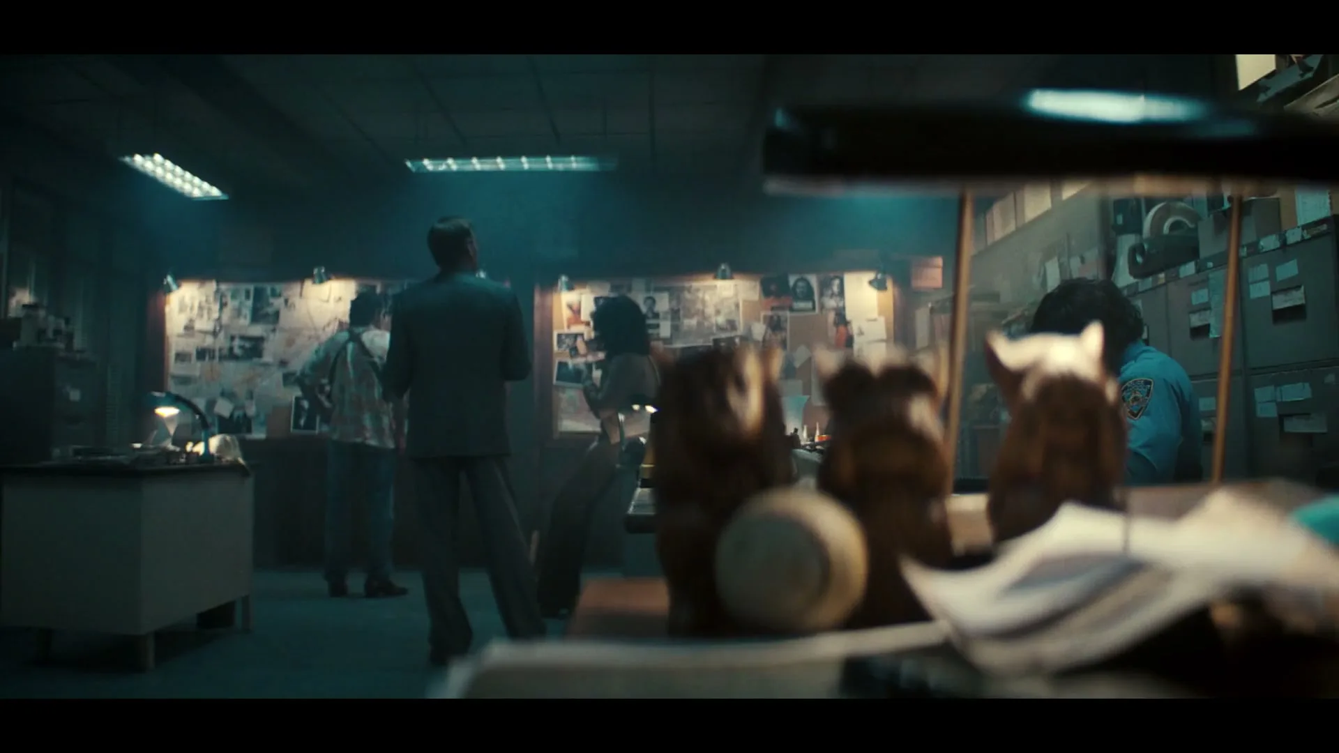 Three wooden monkey statues and a baseball sit on a desk in the foreground, while detectives work in a dimly lit office in the background.