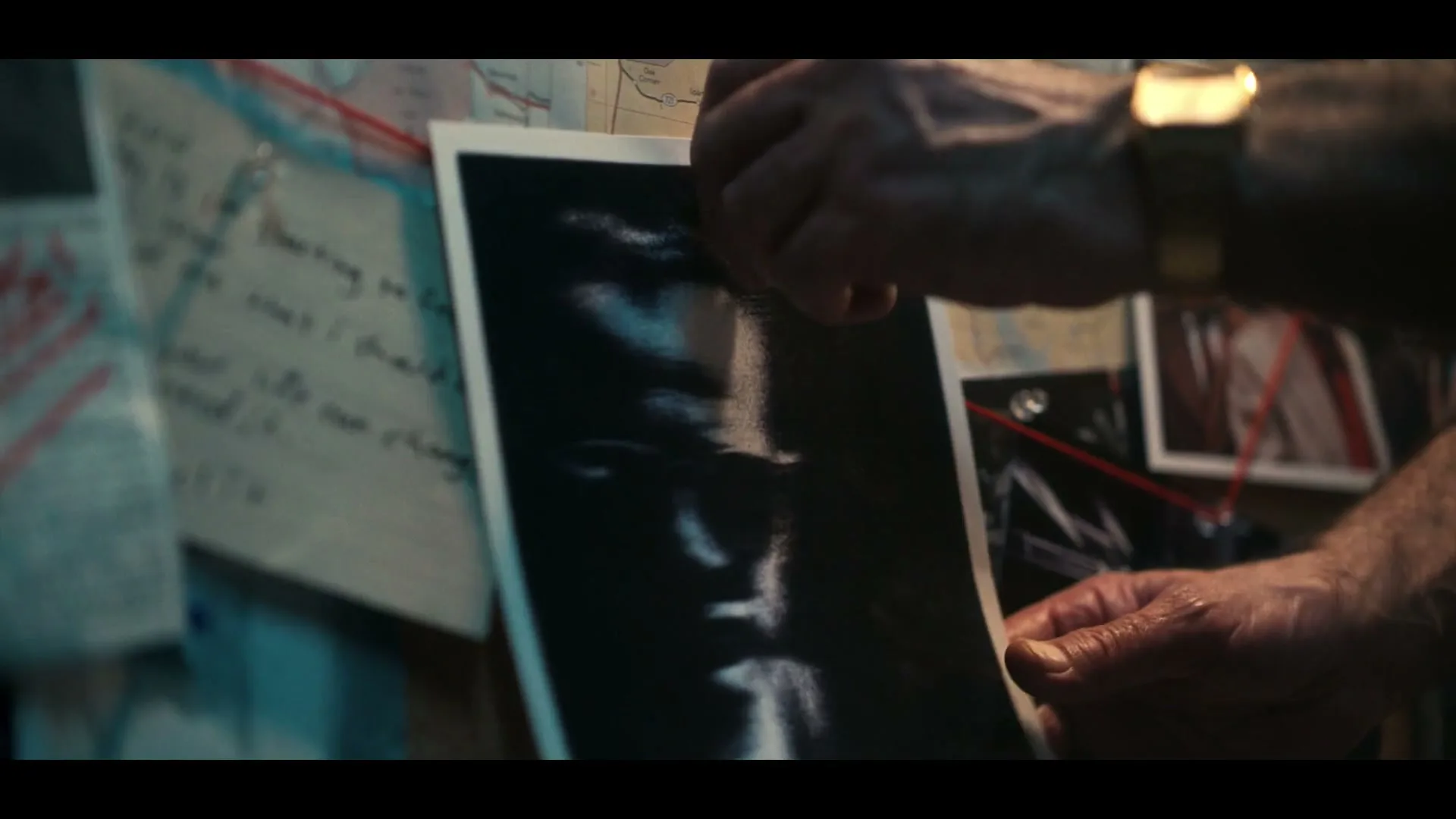 A man's hands pin a black and white photo of a person's face to a cork board covered with maps, notes, and other photos connected by red string.