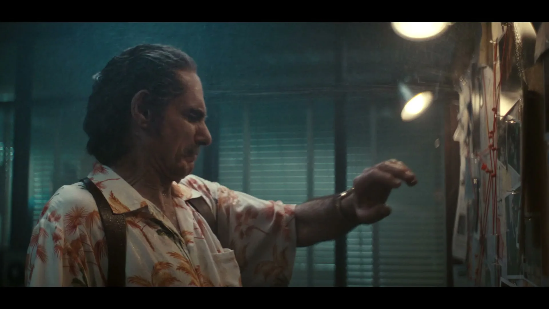 A man with a mustache, wearing a Hawaiian shirt and suspenders, is soaking wet and looking at a cork board with photos and string in a dimly lit office.