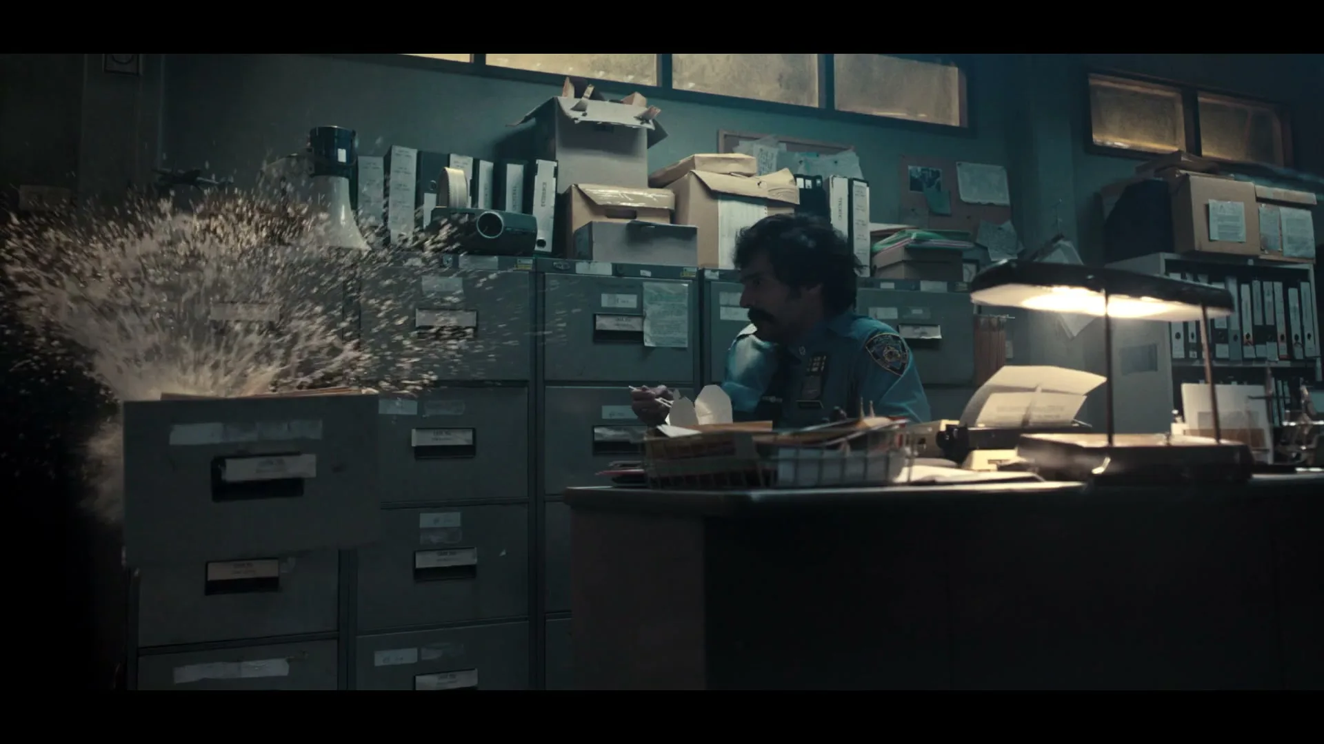 A male detective in a police uniform sits at his desk, eating takeout, as water suddenly explodes from a filing cabinet in a dimly lit office.