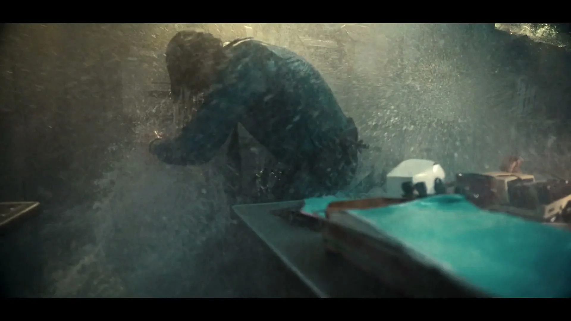 A man in a blue jacket is drenched by a powerful stream of water, struggling to stand in a chaotic, flooded room.