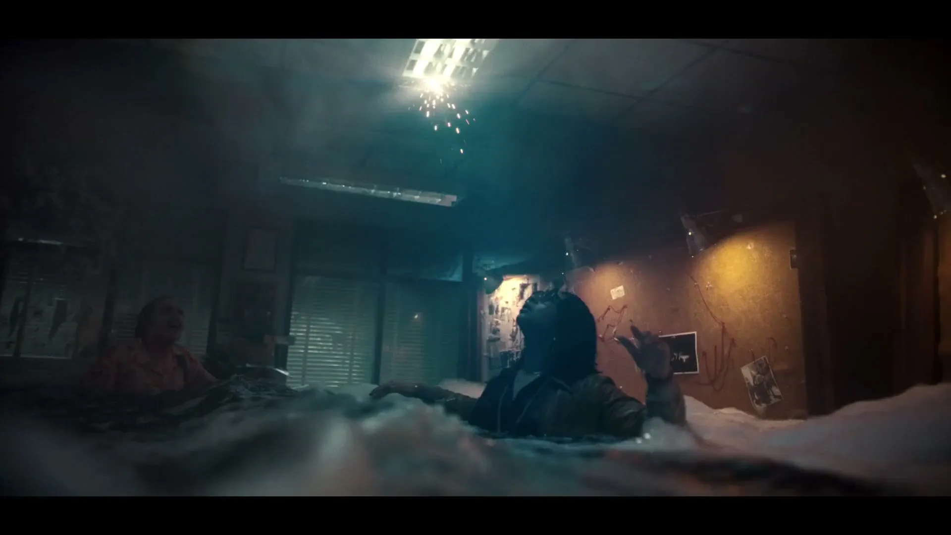 A woman and a man are submerged in water and foam, looking up as sparks fall from a flickering fluorescent light in a flooded office.