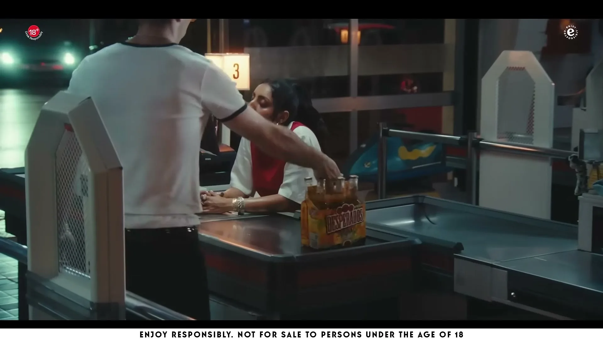 A male customer in a white t-shirt places a pack of Desperados beer on a supermarket checkout counter, while a female cashier in a red and white uniform looks at him.