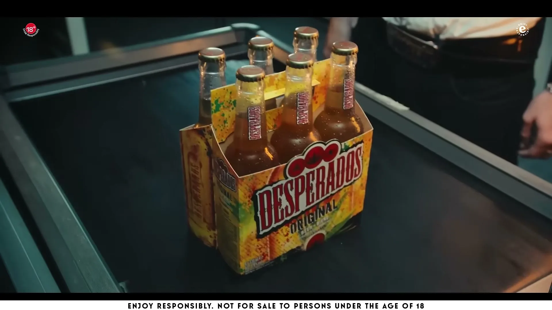 A six-pack of Desperados beer bottles moves along a black conveyor belt at a checkout counter.