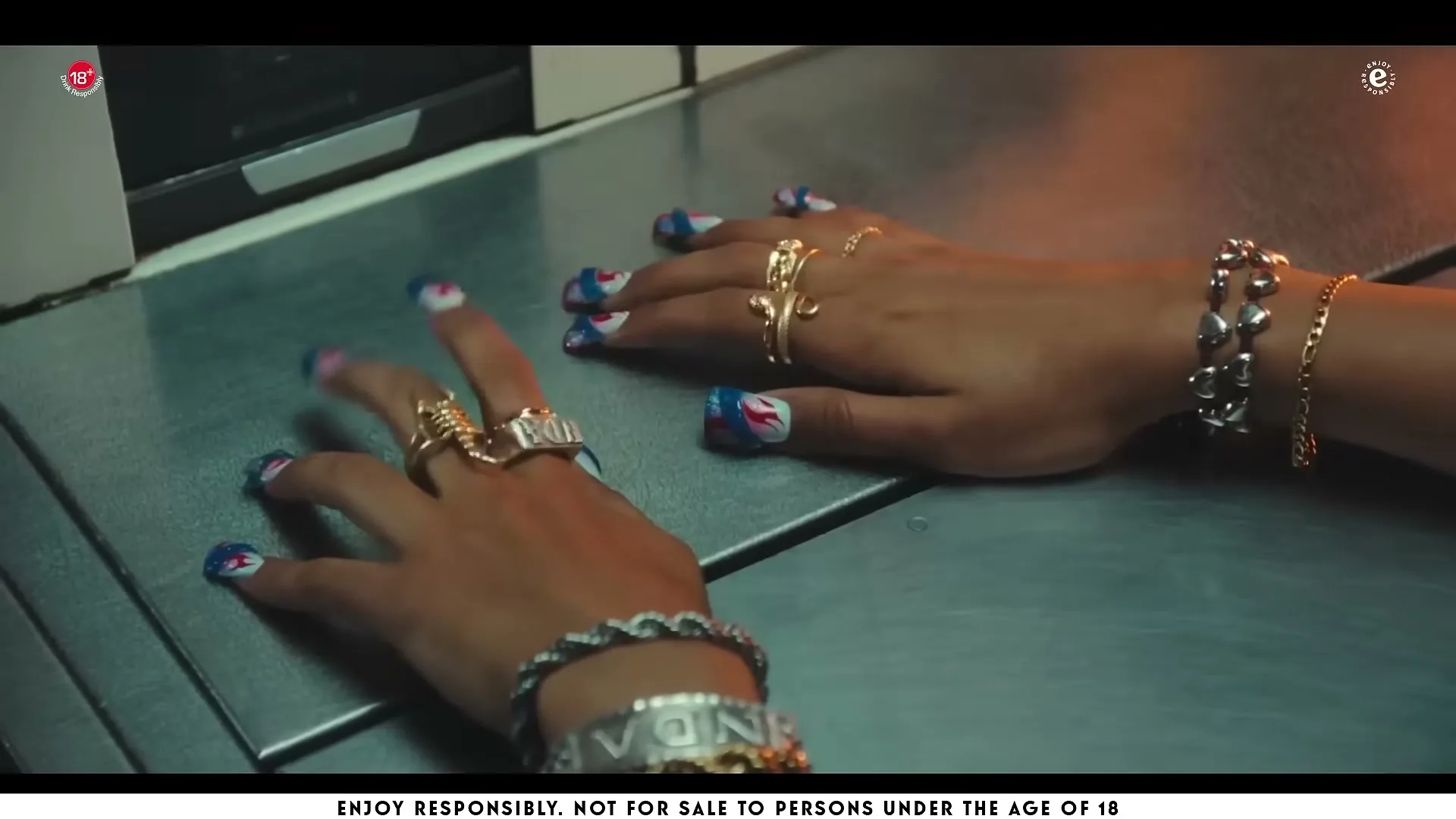 A person's hands with elaborate rings and painted nails rest on a metallic surface in an interior setting.