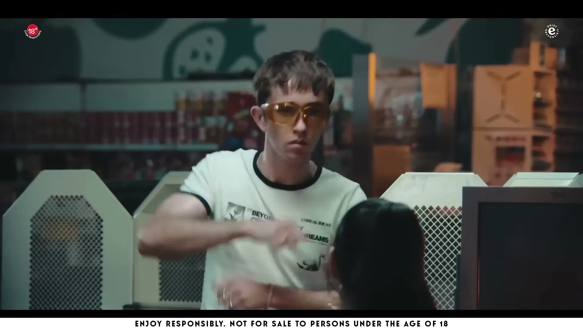 A young man in a white t-shirt and yellow sunglasses gestures with his hands at a checkout counter in a supermarket.