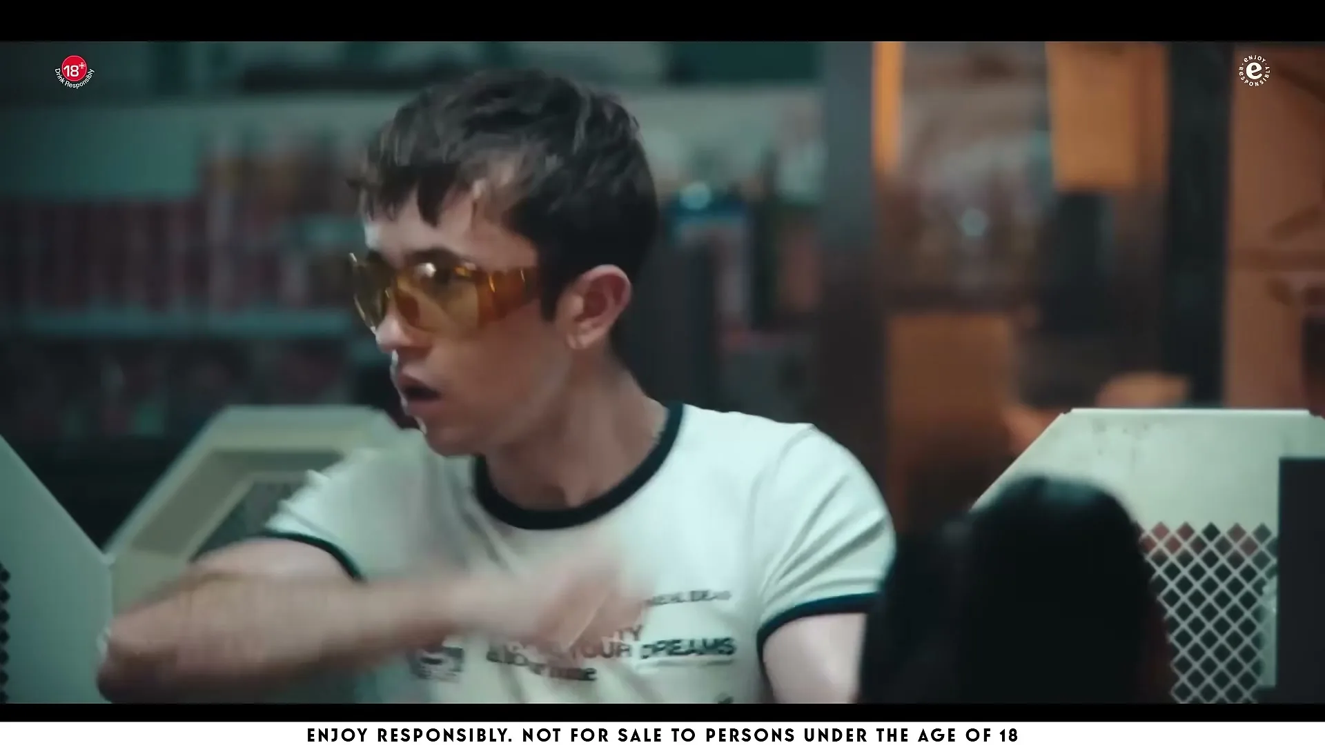 A young man in yellow sunglasses and a white t-shirt with black trim looks surprised and then quickly turns his head to the right in a dimly lit interior.
