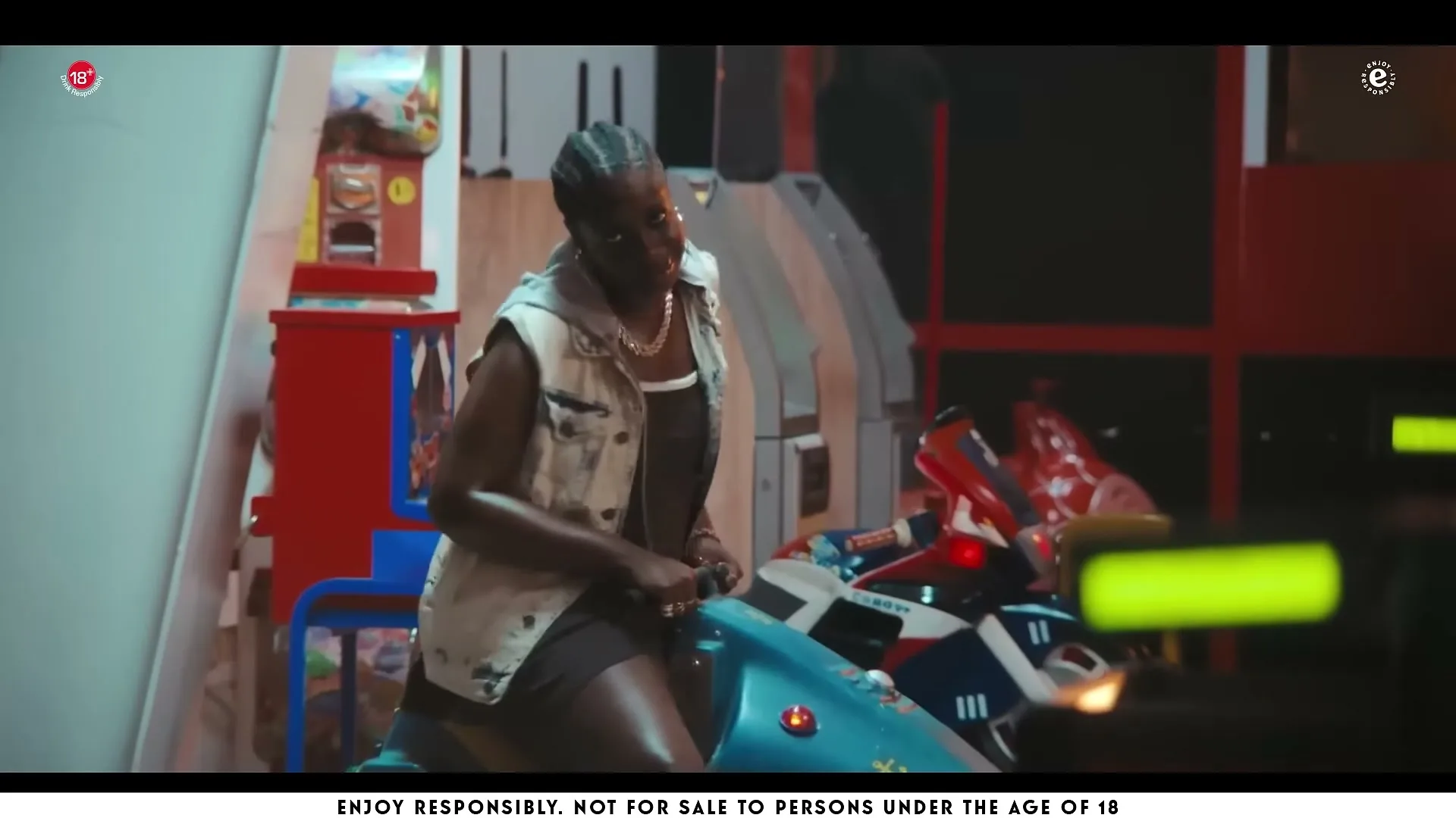 A young woman with braided hair, wearing a denim vest and dark dress, sits on a blue coin-operated motorcycle ride in an arcade.