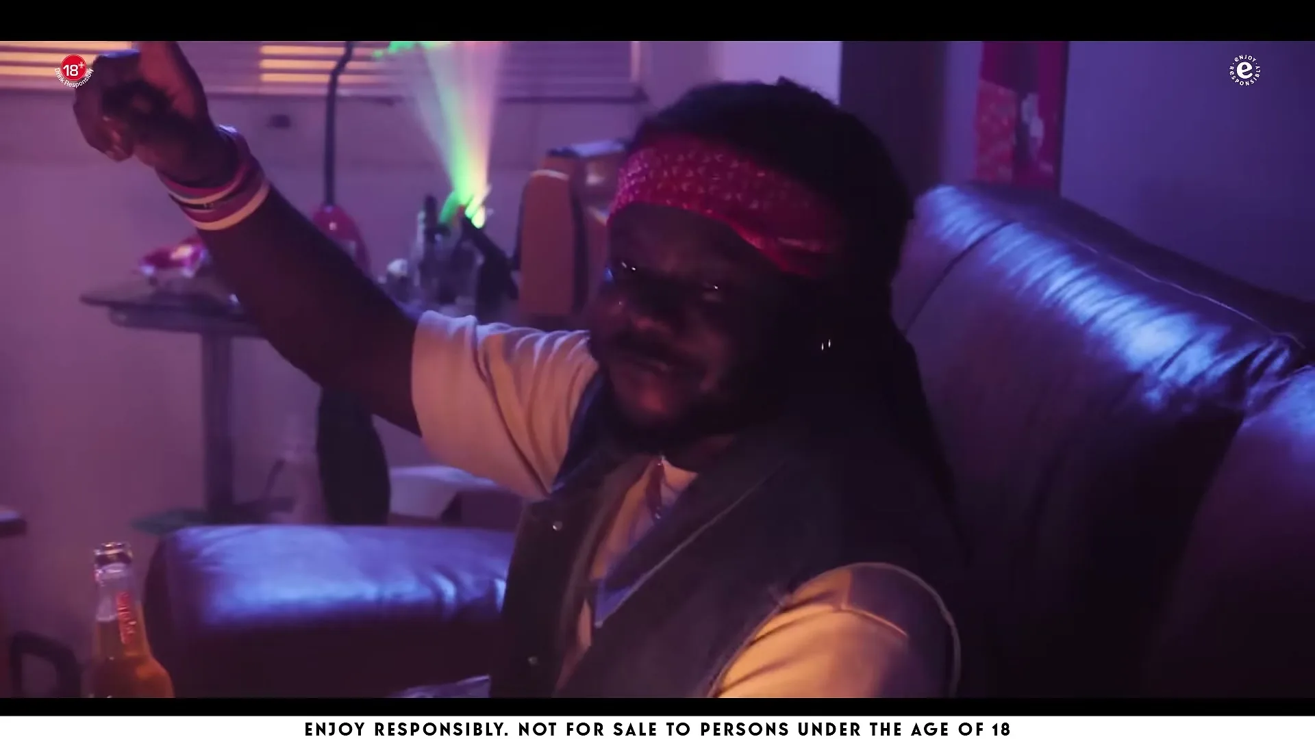 A man with dreadlocks and a red bandana sits on a couch, holding a beer and pointing upwards in a dimly lit room.
