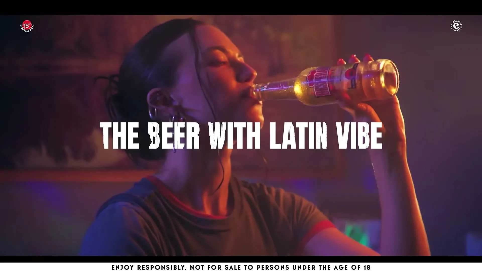 A young woman with dark hair in a casual t-shirt drinks from a Desperados beer bottle in a dimly lit, vibrant interior setting.