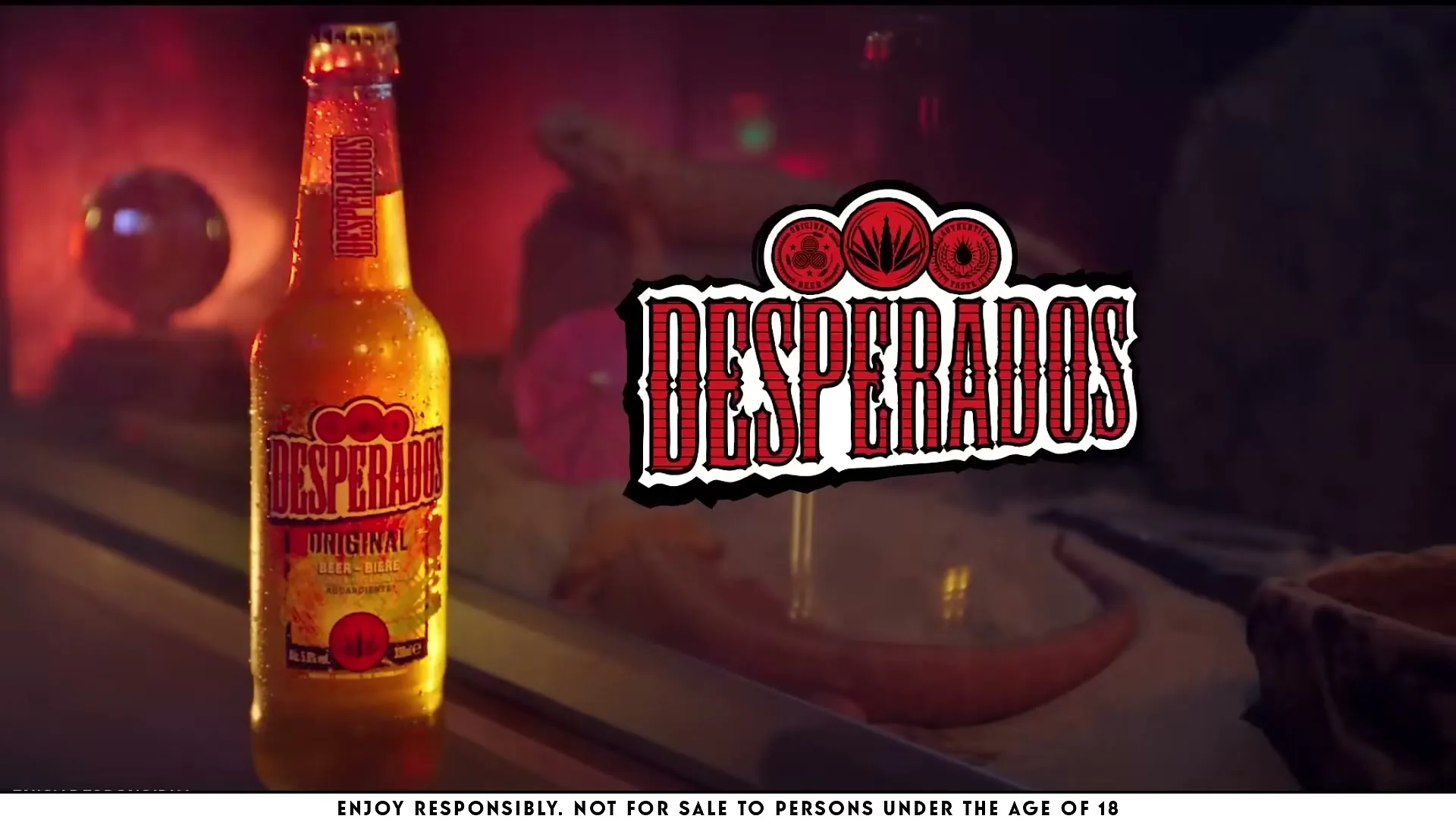 A bottle of Desperados beer stands on a bar counter, with the brand's logo prominently displayed in the background, in a dimly lit interior.