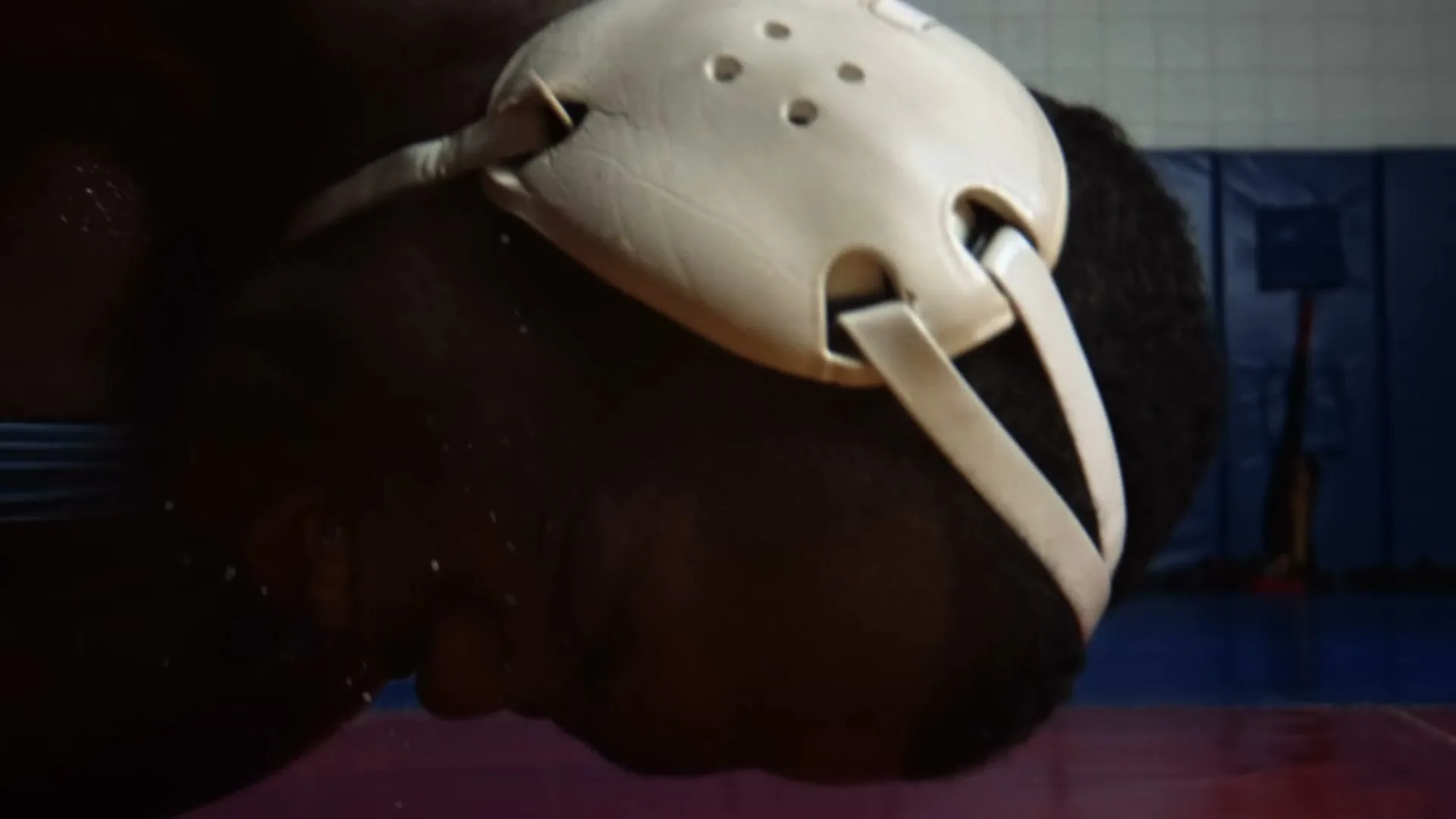 A male wrestler with dark skin and a white headgear is seen from a low angle, sweating and breathing heavily in a wrestling gym.