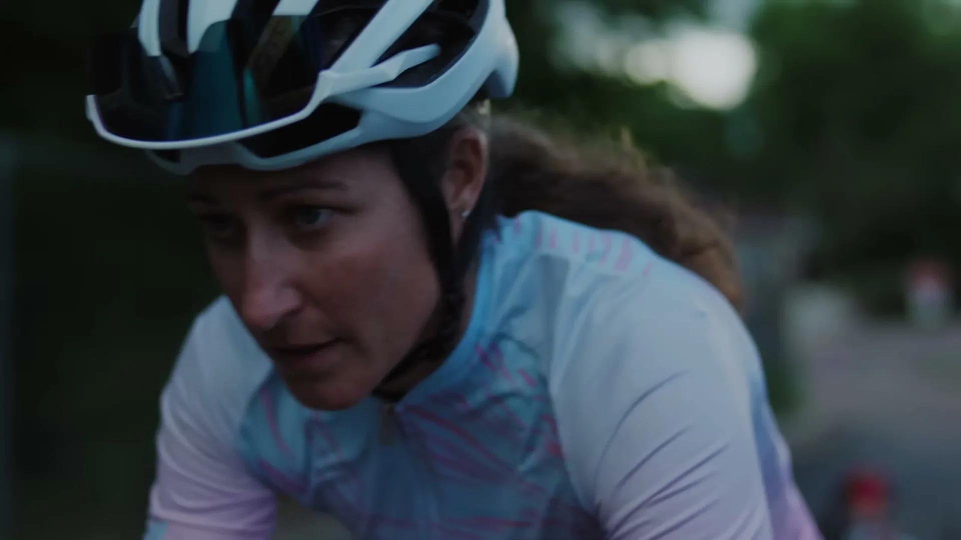 A female cyclist, wearing a helmet and a light blue and pink jersey, looks down with a determined expression while riding outdoors.