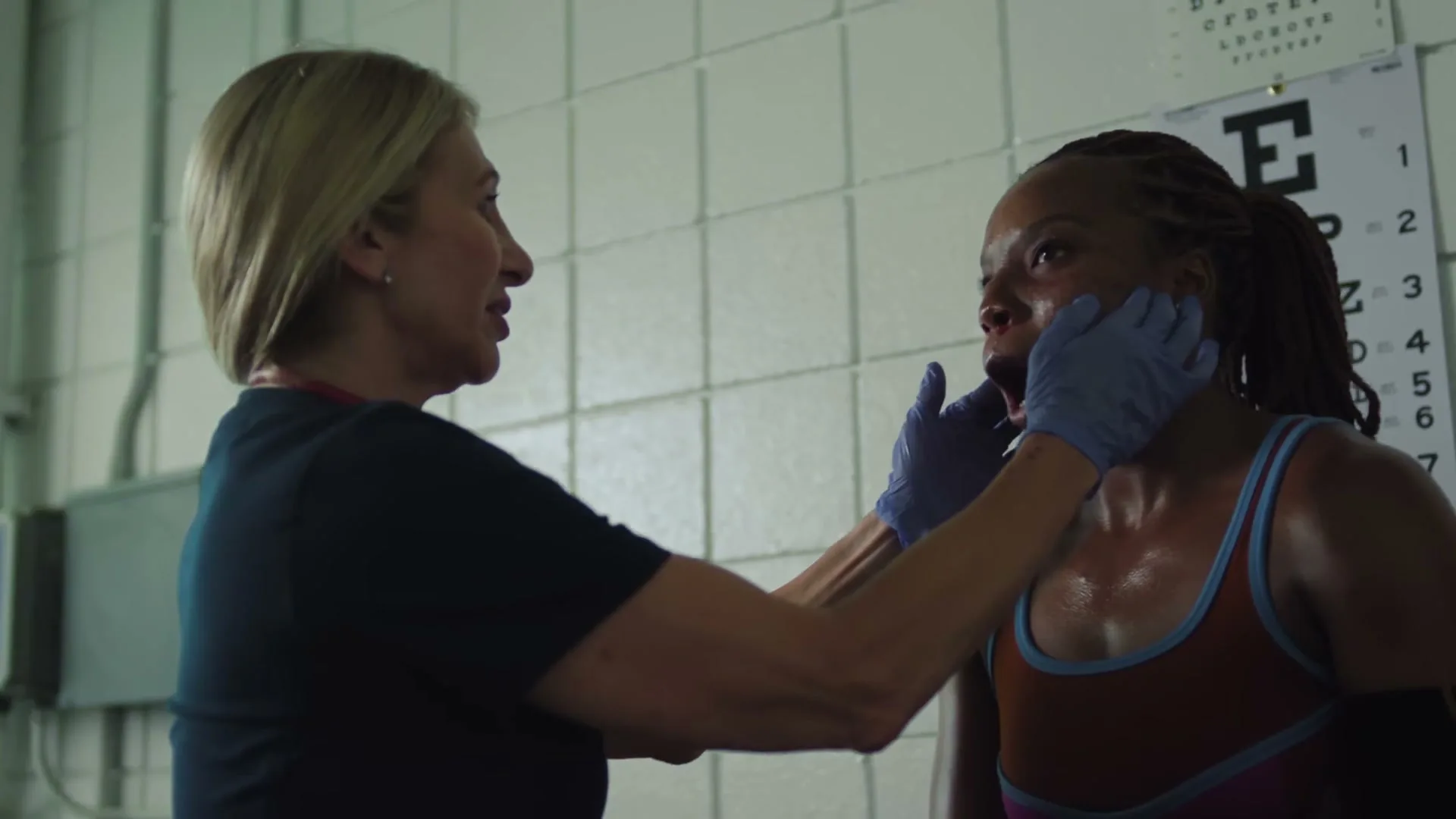 A female medical professional in scrubs and gloves examines a sweaty female boxer's face in a locker room with an eye chart on the wall.