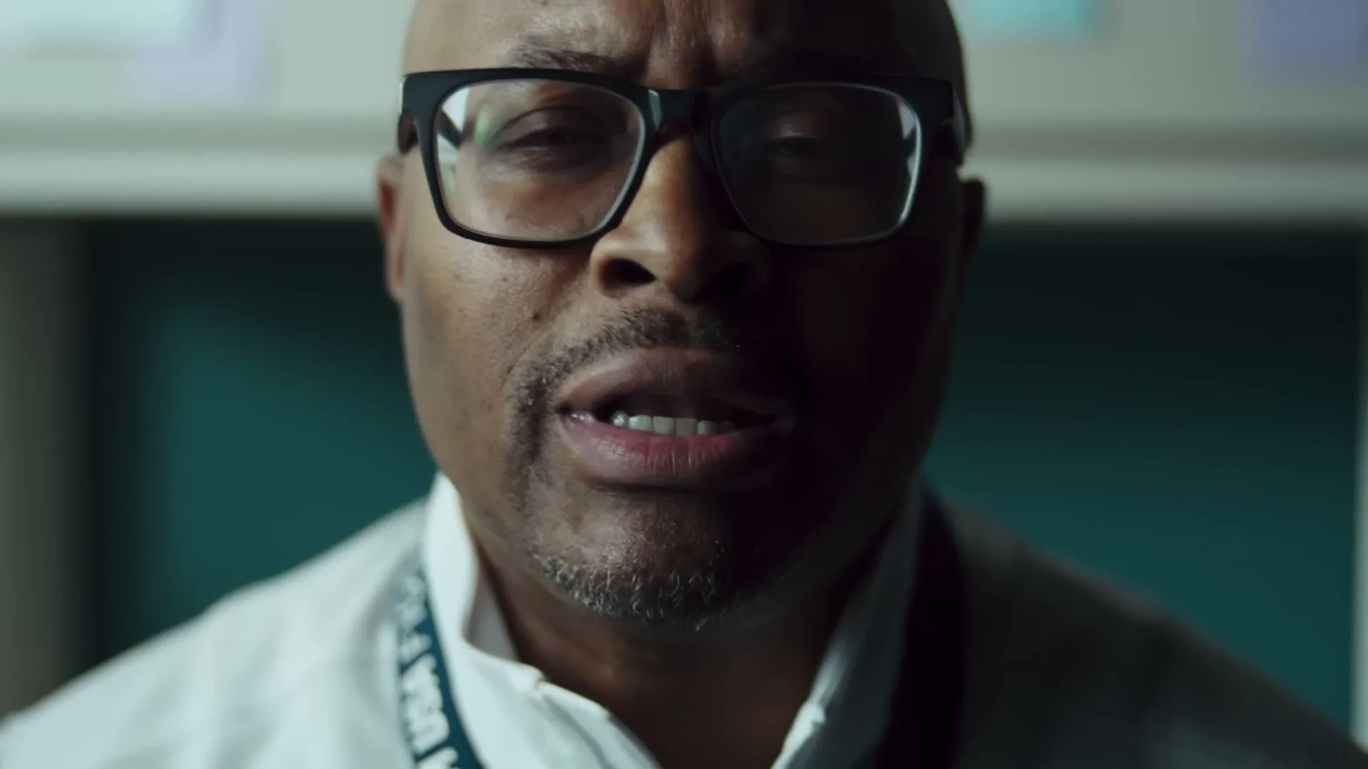 A bald African American man with glasses and a goatee, wearing a white lab coat, opens his eyes and speaks in an interior setting.